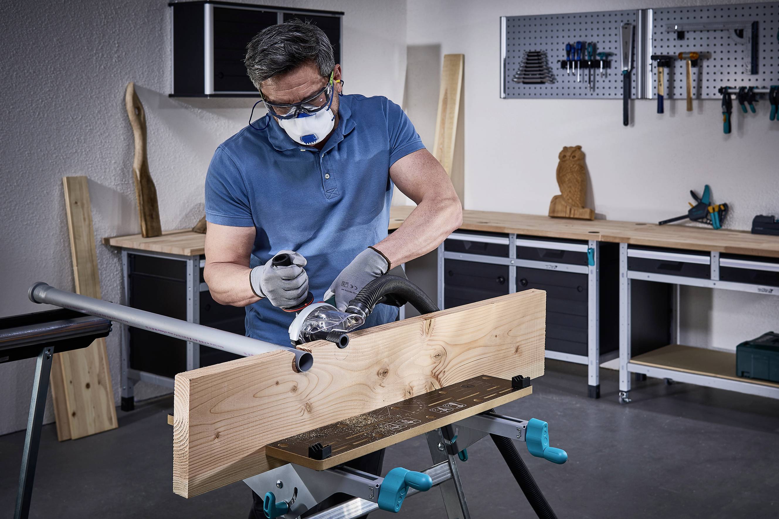 Mężczyzna obrabia deskę drewnianą elektronarzędziem w warsztacie, nosi maskę ochronną i okulary. W tle widoczne są narzędzia.