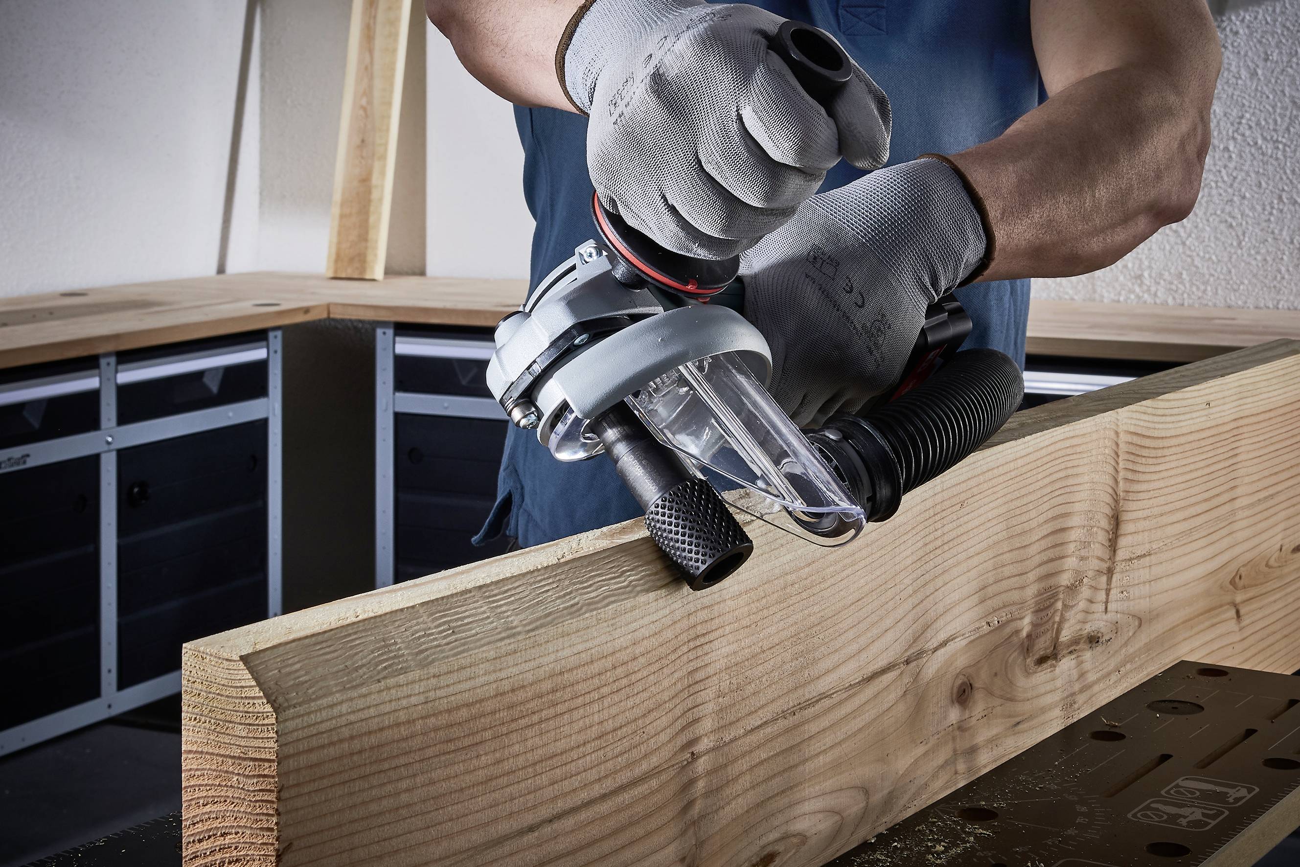 Osoba szlifuje krawędź drewna elektroniczną ręczną szlifierką. Nosi rękawice robocze, a otoczenie stanowi warsztat.