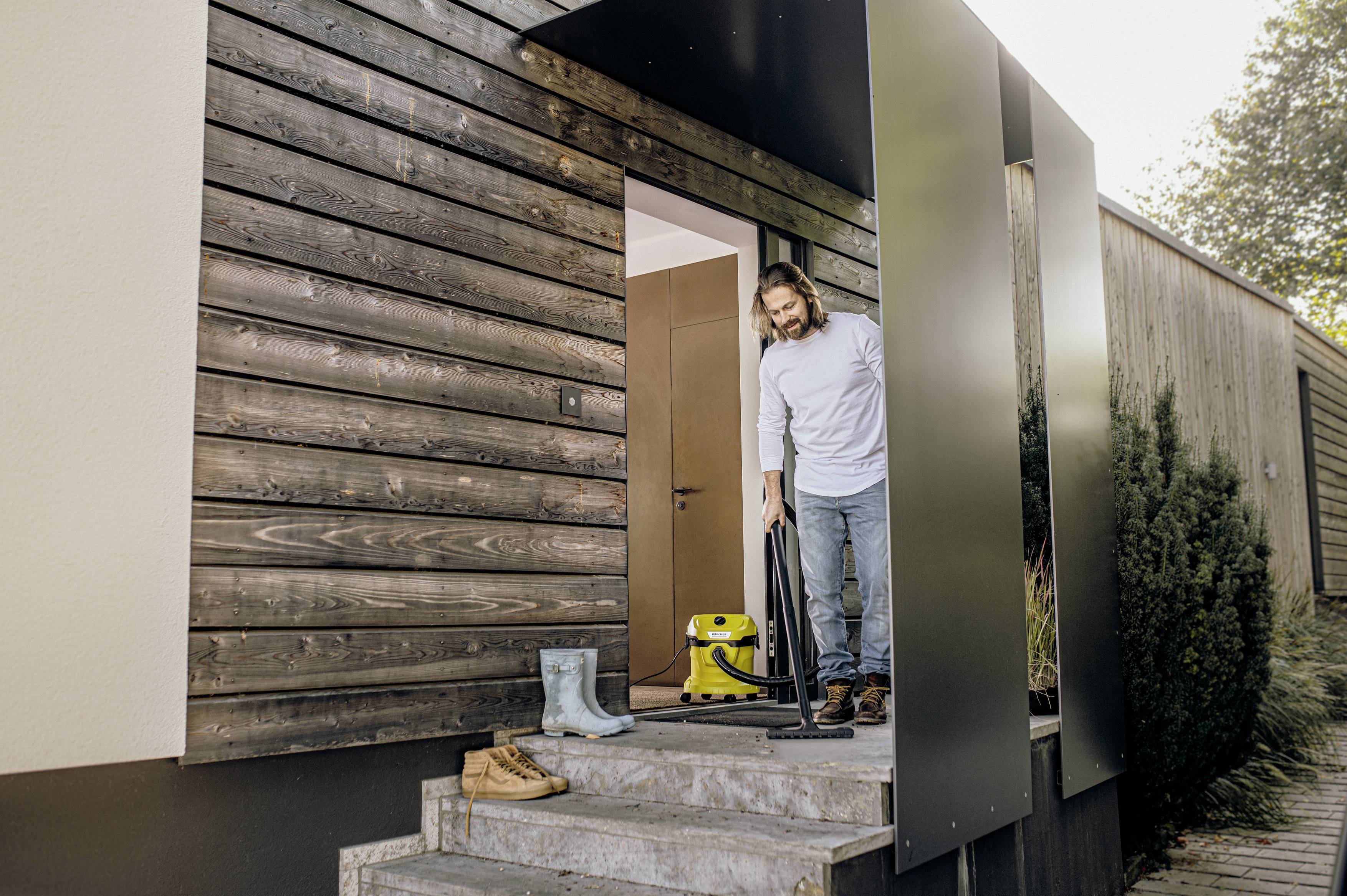 Osoba stoi na schodach domu i odkurza wycieraczkę za pomocą odkurzacza. Na stopniach stoją kalosze i para butów.