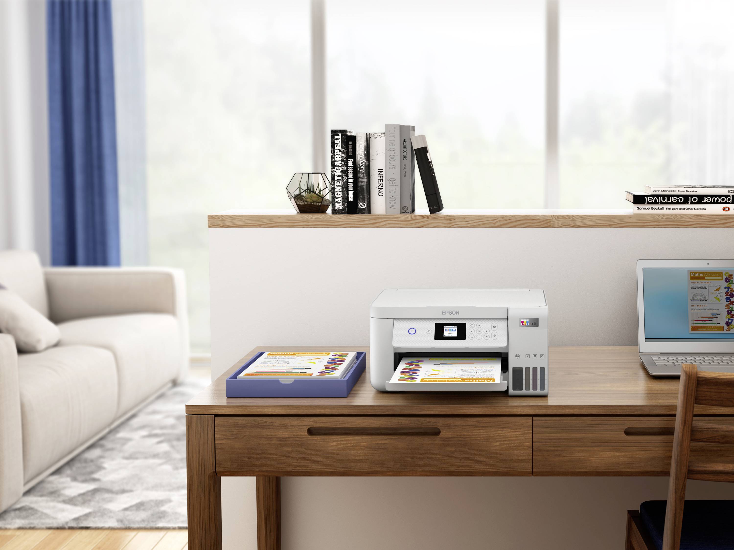 Drukarka na drewnianym stole drukuje kolorowy dokument; obok leży laptop i broszura. W tle widoczny jest salon z kanapą i regałem na książki.