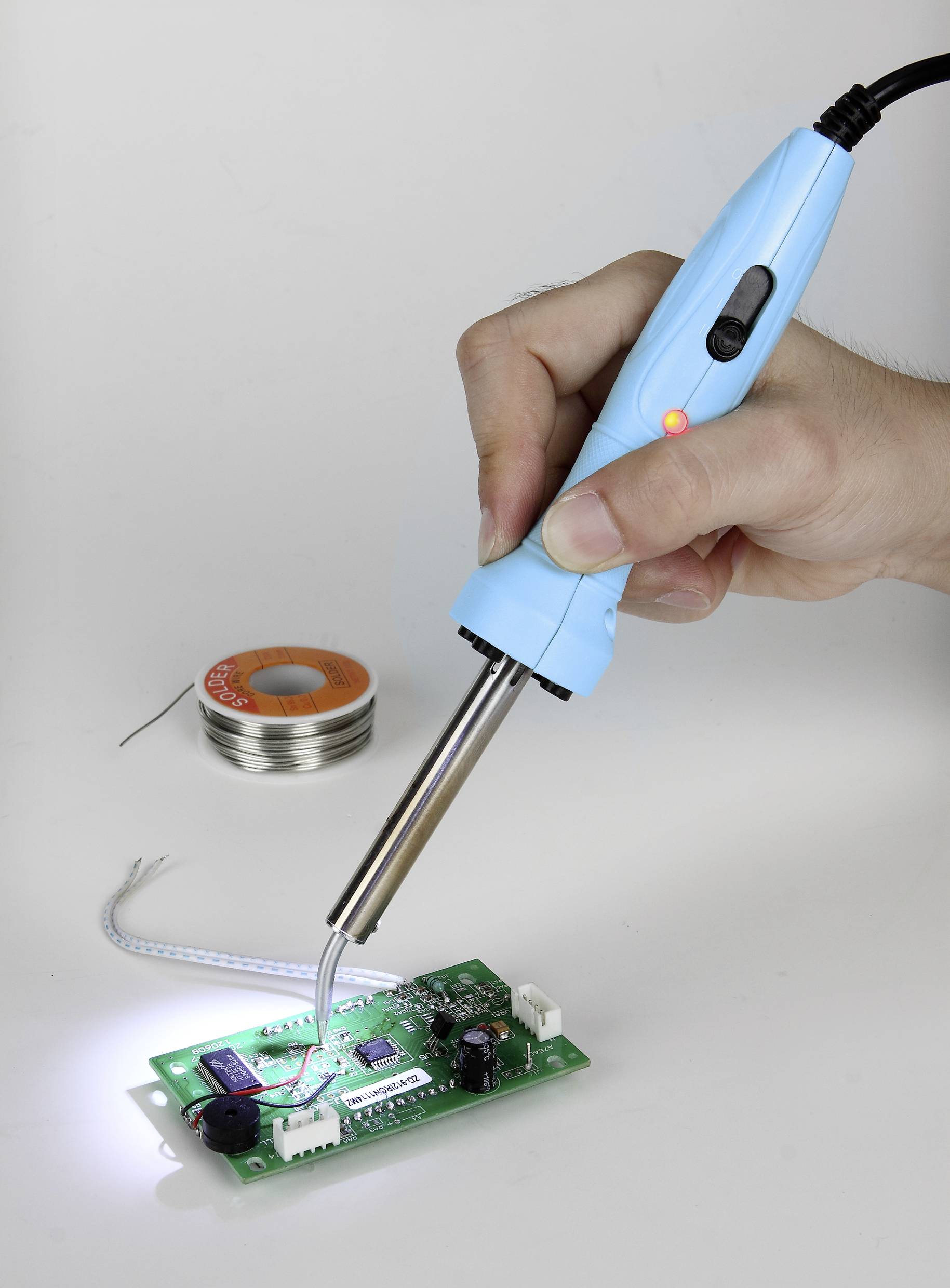 Osoba trzyma lutownicę i lutuje przewody na zieloną płytkę drukowaną. W tle widać rolkę lutu.