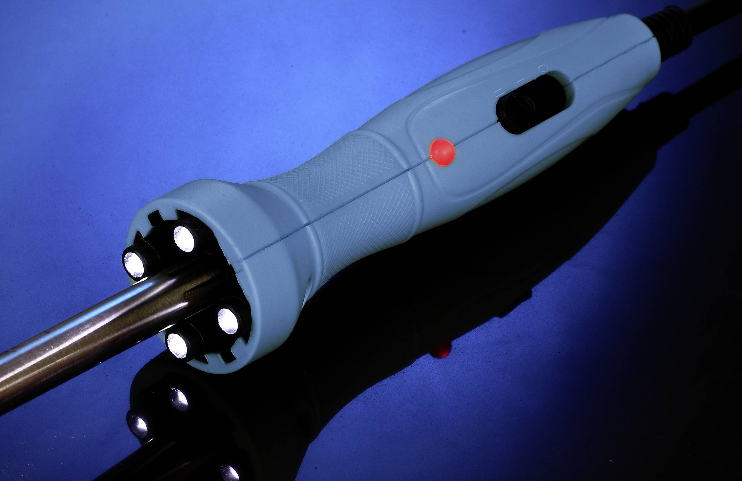 Lutownica z niebieskim uchwytem leży na powierzchni odbijającej światło na niebieskim tle.