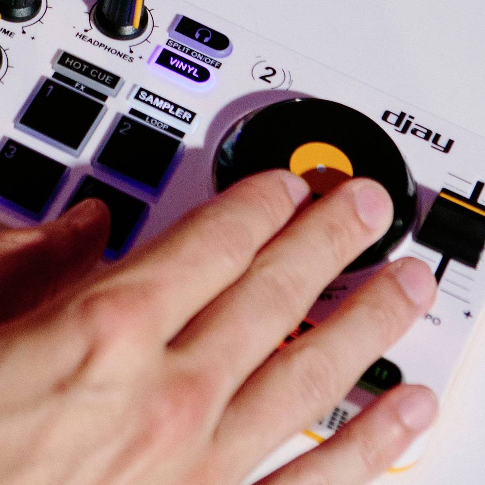 Ręka obsługuje kontroler DJ-ski z wieloma przyciskami i dużym pokrętłem, które jest używane do miksowania muzyki.