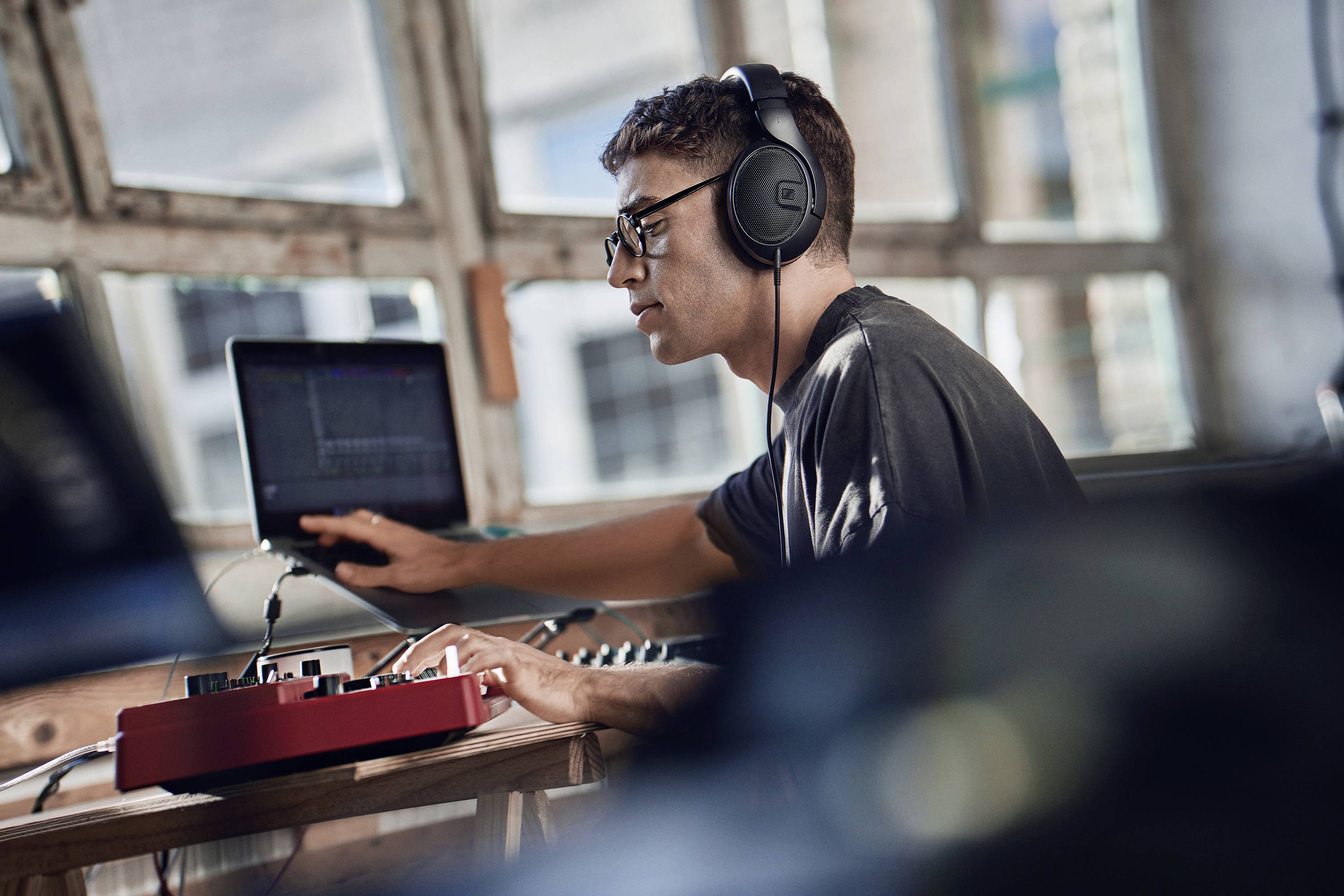 Młody mężczyzna w słuchawkach pracuje przy laptopie i urządzeniu muzycznym. Jest skoncentrowany w jasnym pomieszczeniu z wieloma oknami.