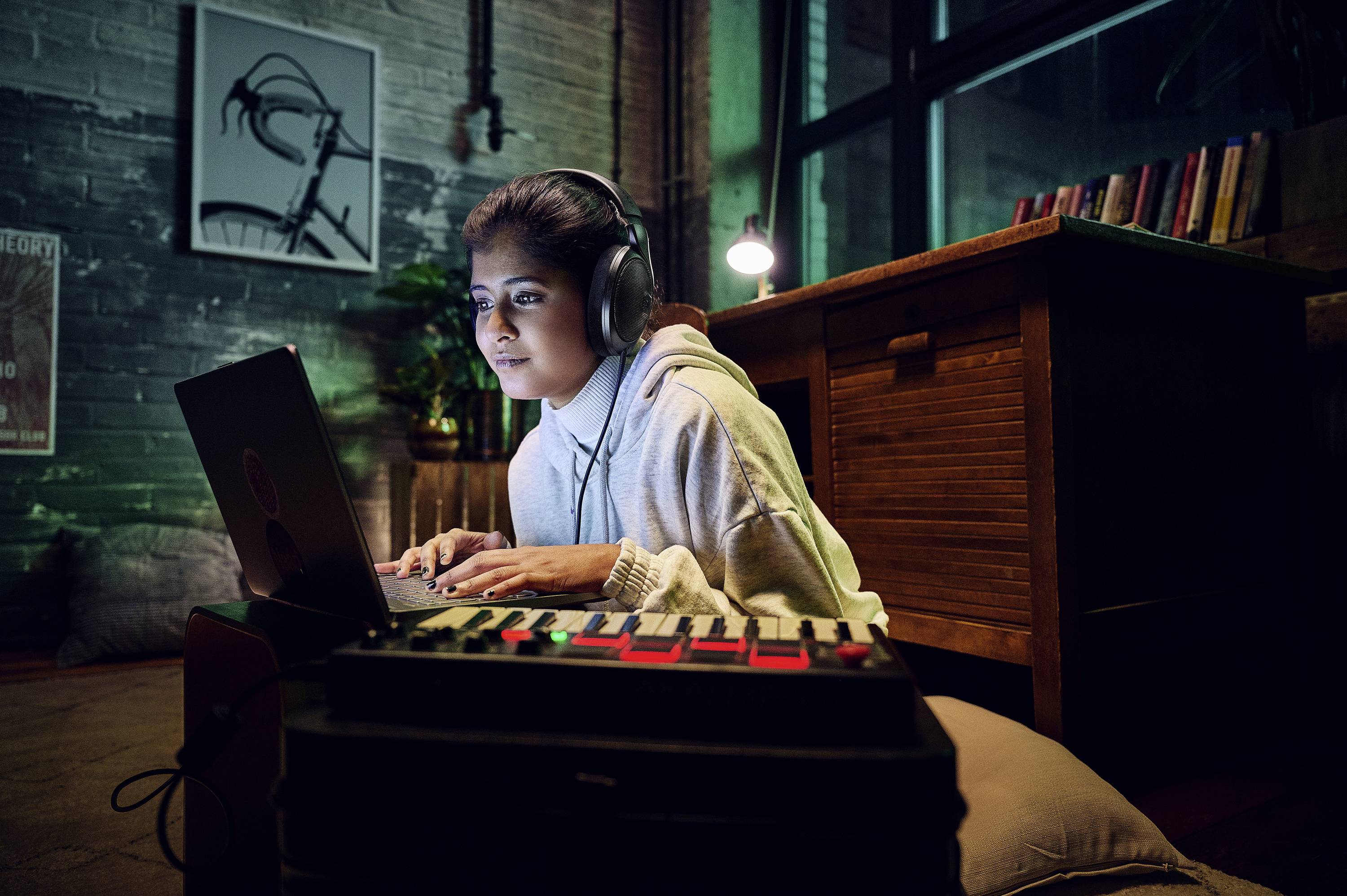 Osoba w słuchawkach pracuje skupiona przy laptopie w przytulnym pokoju o stonowanym oświetleniu.
