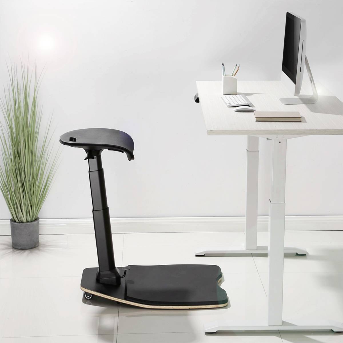 Czarny ergonomiczny fotel na kółkach obok biurka z regulowaną wysokością z komputerem i przyborami biurowymi.