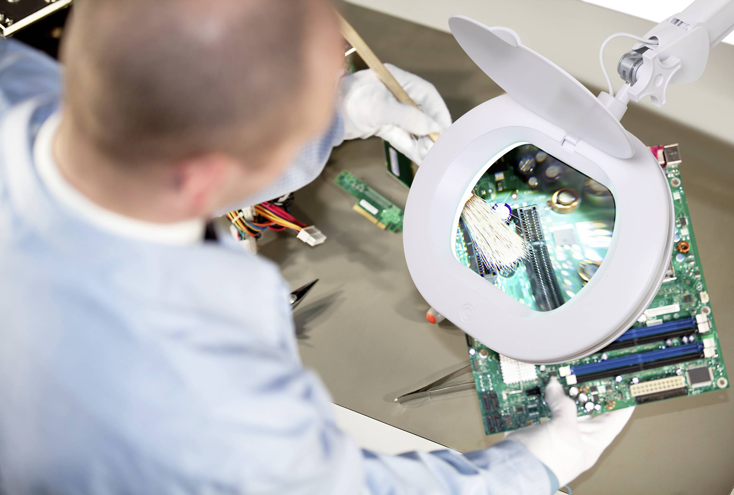 Osoba w stroju ochronnym sprawdza płytkę komputerową za pomocą lampy z lupą, dokładnie kontrolując jej komponenty elektroniczne.