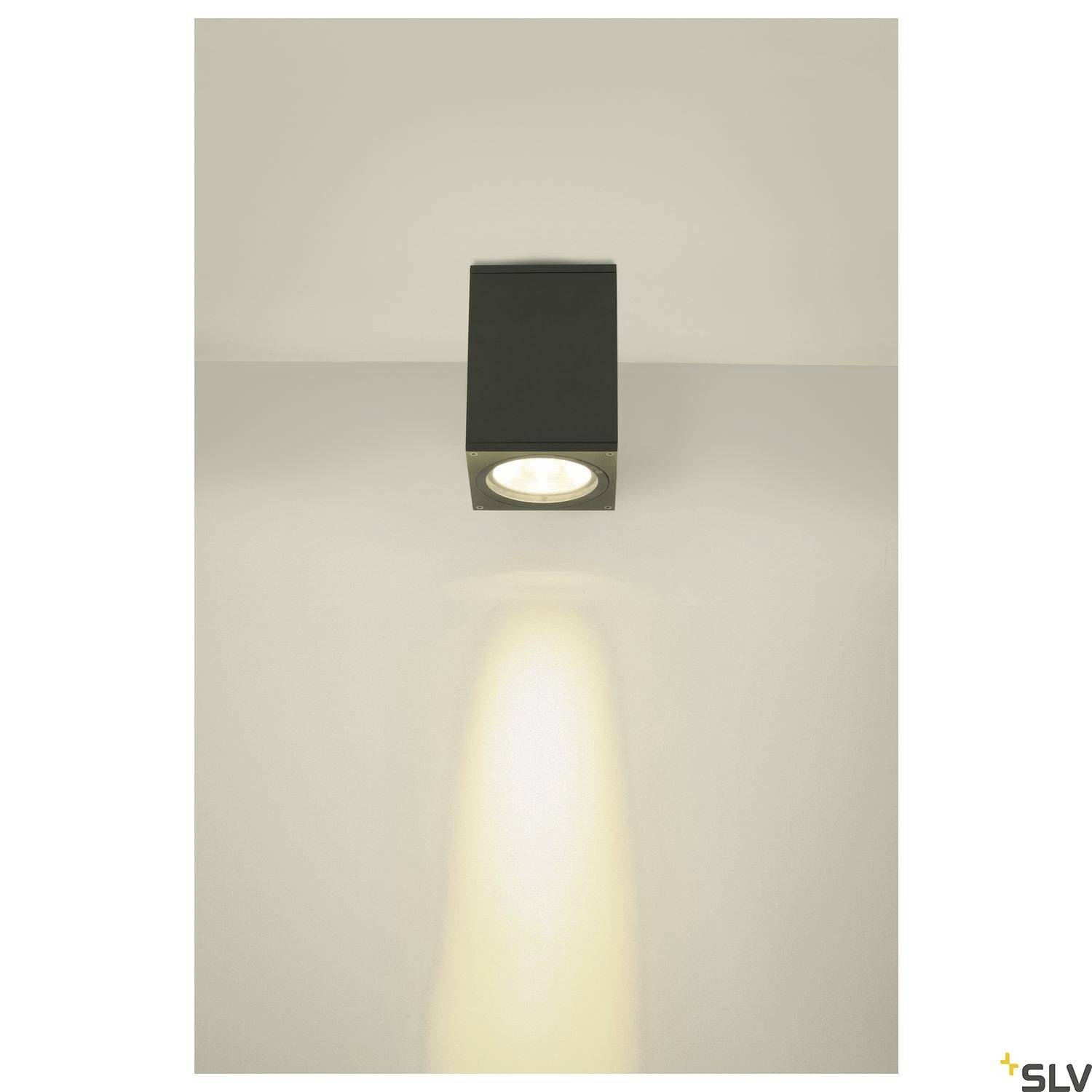 Czarna lampa sufitowa rzuca światło w dół na ścianę lub podłogę. Otoczenie jest urządzone w minimalistycznym stylu.