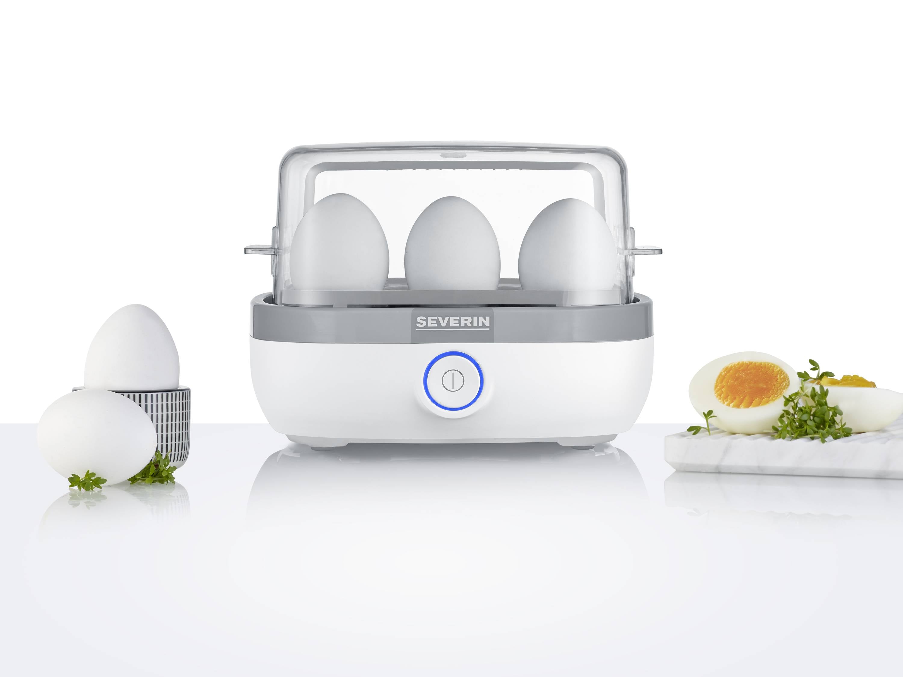 Minutnik do jajek z czterema jajkami w środku oraz ugotowanym, przekrojonym na pół jajkiem na desce. Minutnik jest biały z szarymi akcentami.