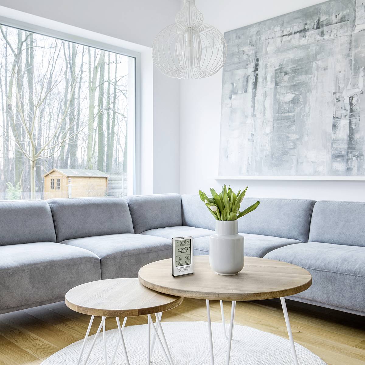 Jasny salon z szarą sofą, drewnianym stolikiem i dużą ścianą okienną. Na zewnątrz widoczne są drzewa, po lewej stronie stoi drewniany pawilon.