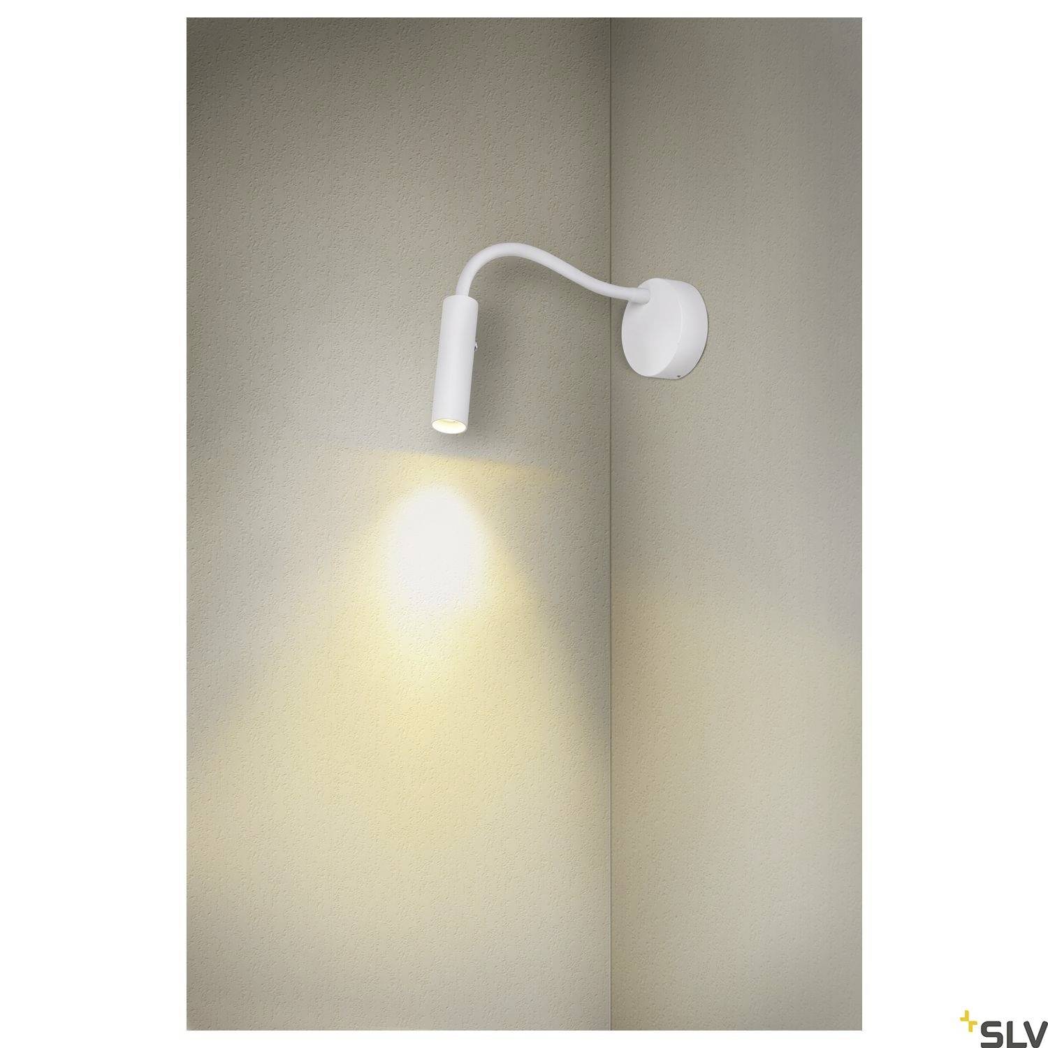 Biała, minimalistyczna lampa ścienna z giętym ramieniem oświetla beżową ścianę. Lampa rzuca ciepłe światło w dół.