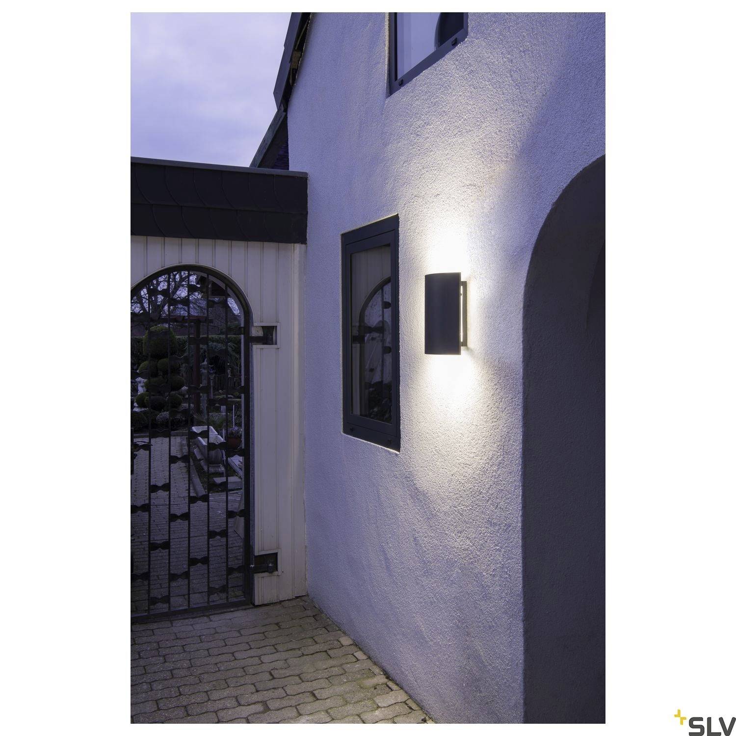 Lampa na ścianie zewnętrznej oświetla fasadę domu o zmierzchu; obok lampy znajdują się drzwi z dekoracyjną metalową kratą.