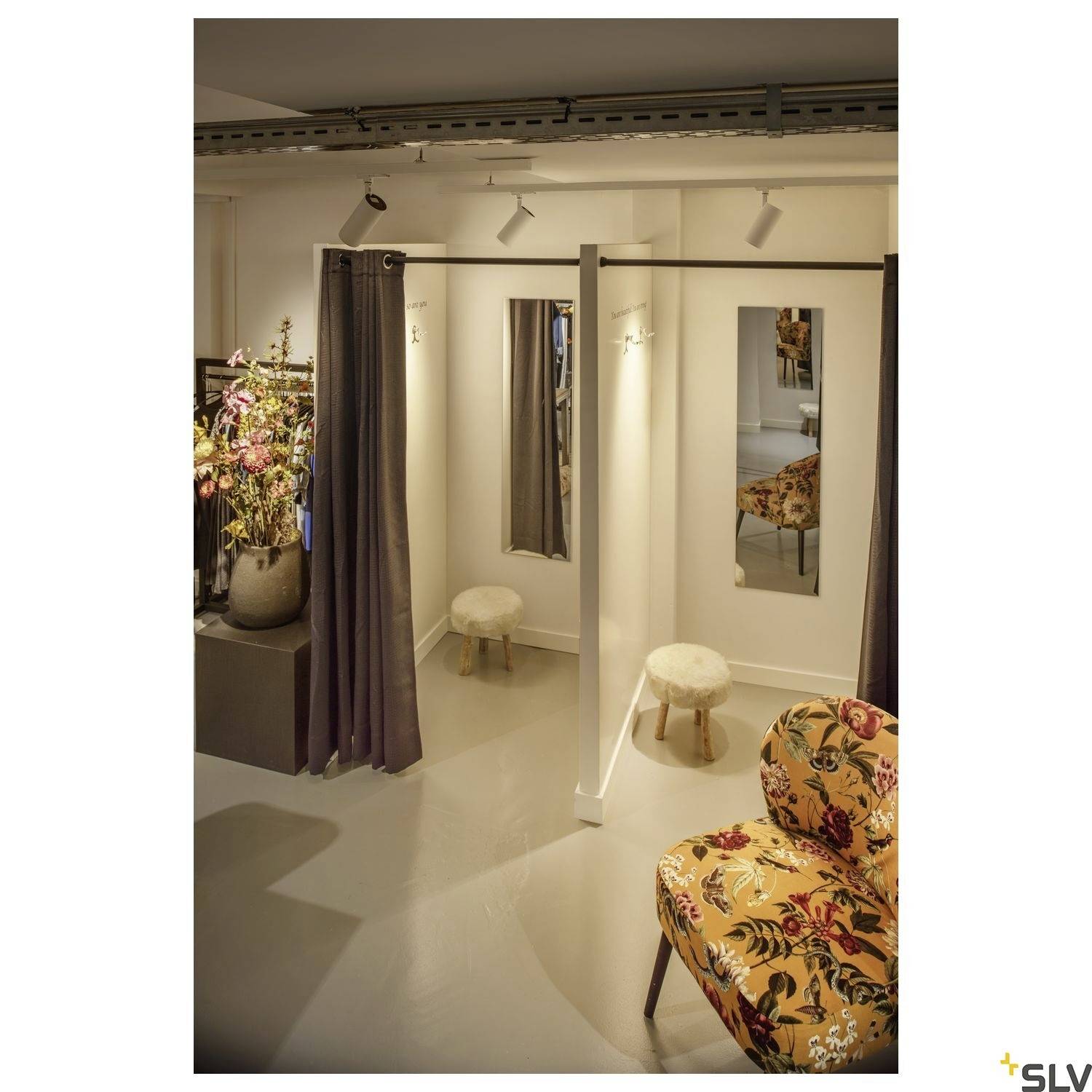 Przebieralnia z dwoma kabiami, lustami i stołkami. Po lewej stronie stoi bukiet kwiatowy, po prawej - tapicerowany fotel we wzory kwiatowe.