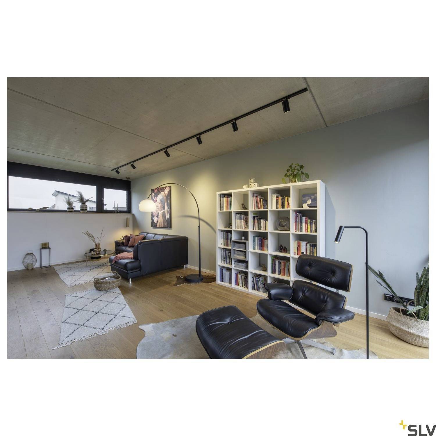 Nowoczesny salon z niebieskimi ścianami, drewnianą podłogą, białą biblioteką, czarnym fotelem i lampą stojącą. Światło wpadające przez duże okno.