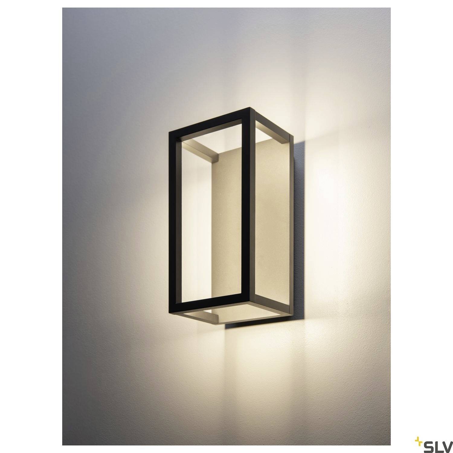 Kinkiet z prostokątną metalową ramą, emitujący ciepłe, rozproszone światło na białą ścianę.