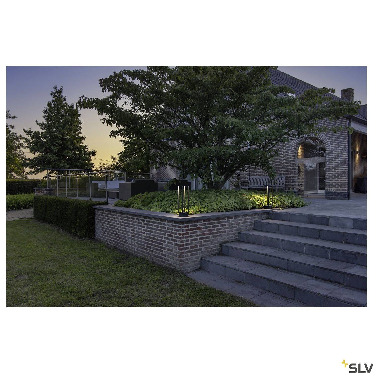 Obraz przedstawia zadbany ogród z oświetlonymi schodami i dużym drzewem przed murowanym domem o zmierzchu.