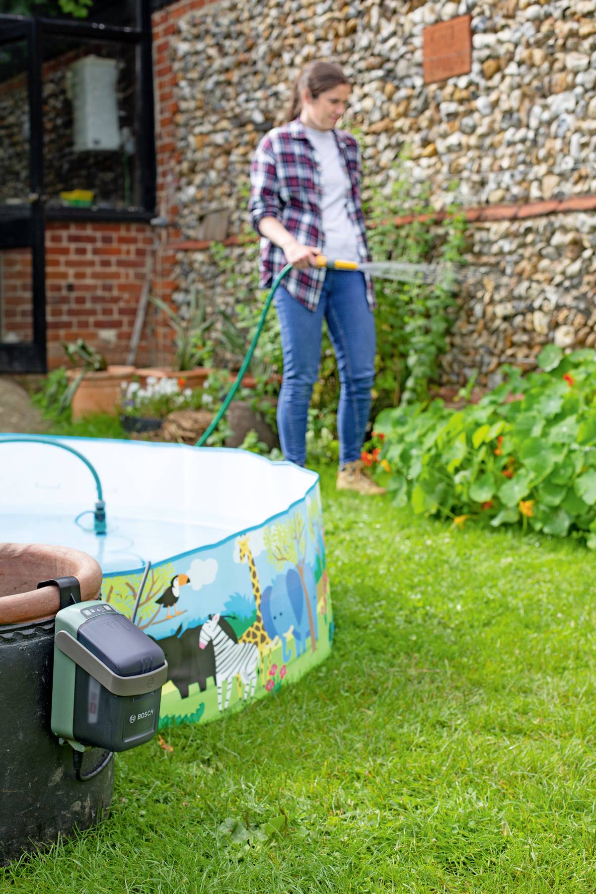 Osoba w ogrodzie podlewa rośliny wężem ogrodowym. Na trawniku stoi kolorowo ozdobiony basen dla dzieci.