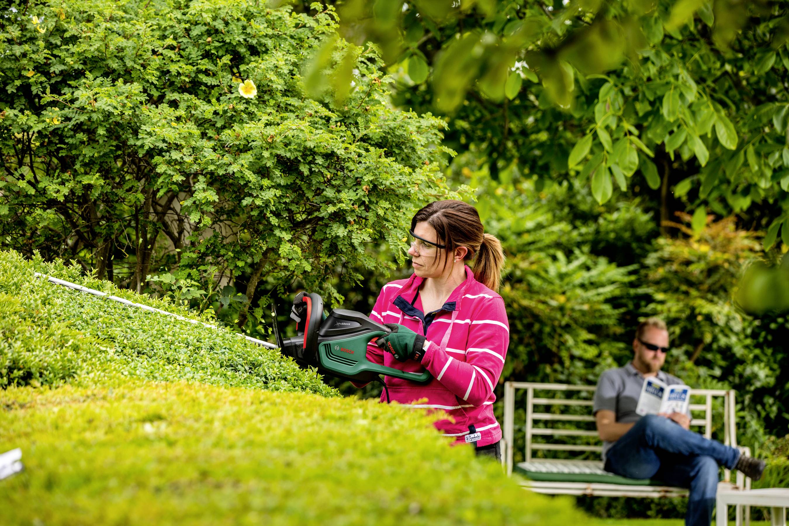 Kobieta przycina żywopłot elektrycznym nożycami do żywopłotu, podczas gdy w tle mężczyzna siedzi na ławce i czyta książkę.
