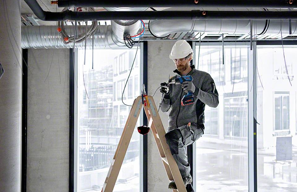Robotnik budowlany stoi na drabinie i używa akumulatorowego wiertarko-wkrętarki do instalacji przewodów elektrycznych na suficie nieukończonego budynku.