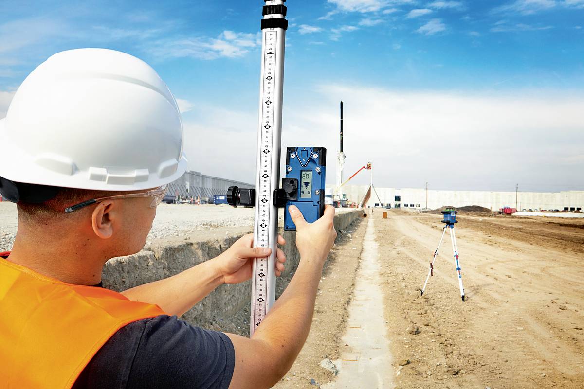 Robotnik budowlany w kasku i kamizelce mierzy teren za pomocą niwelatora na placu budowy. W tle widoczne są prace budowlane.