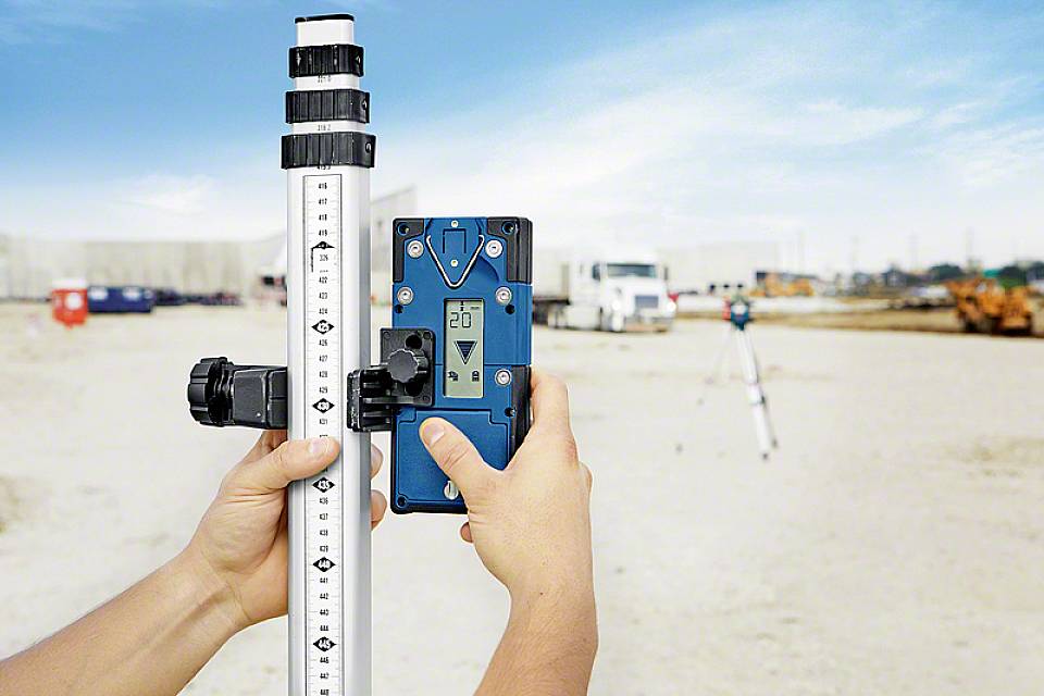 Dwie ręce trzymają cyfrowy przyrząd pomiarowy przed terenem budowy. Mierzona jest duża łata pomiarowa. W tle widoczne są maszyny budowlane.