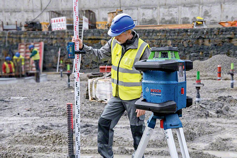 Robotnik budowlany w kasku i kamizelce ochronnej używa laserowego urządzenia pomiarowego na placu budowy, aby określić wysokości.