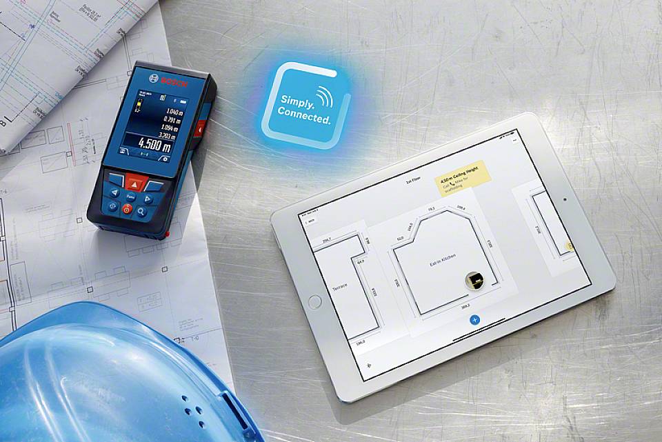 Tablet wyświetla rzut budynku. Obok leży cyfrowy laserowy miernik odległości i plan budowy. Tekst na tablecie: 'Usunąć folię'. Widoczny jest również kask budowlany.