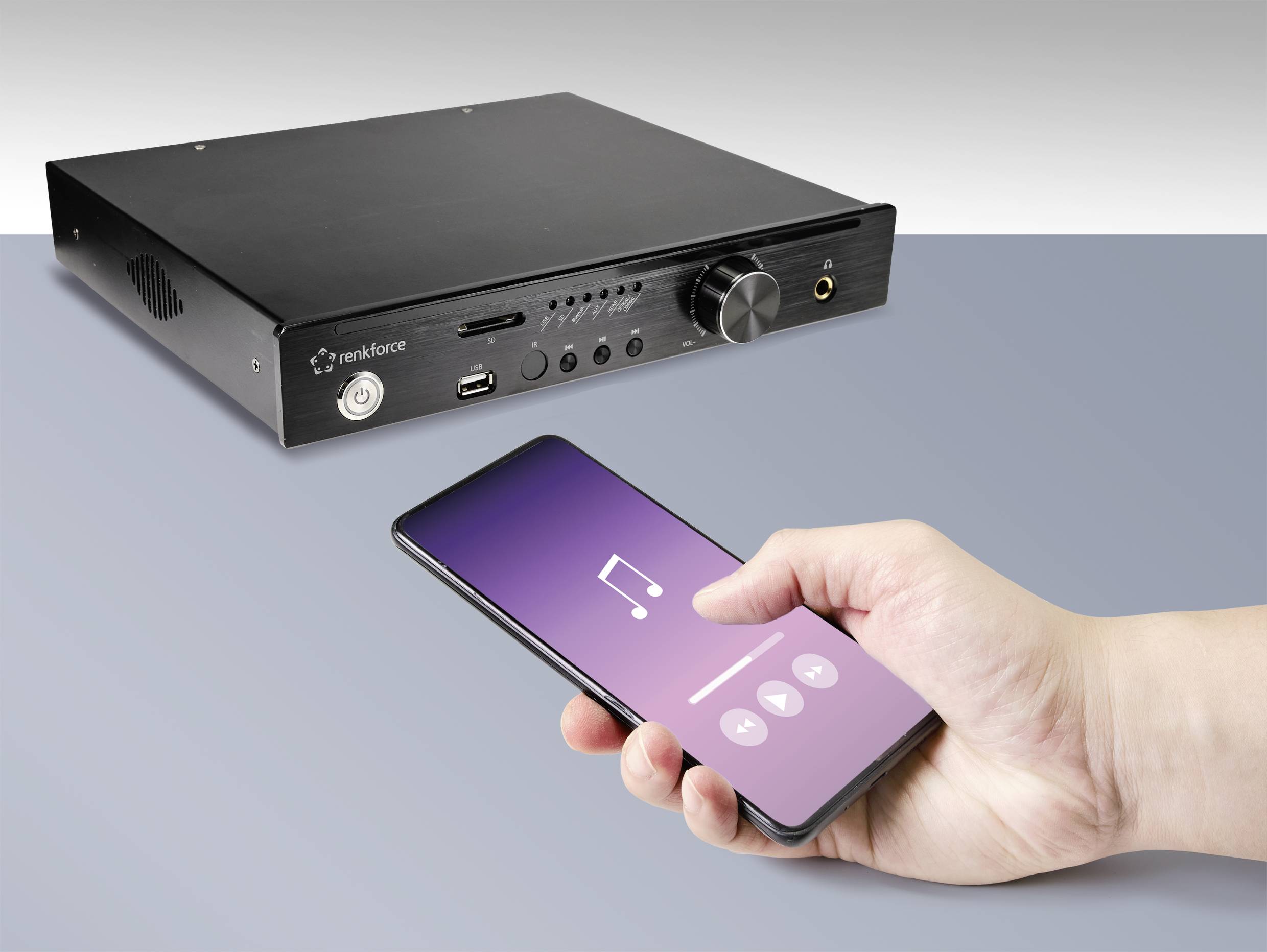 Osoba steruje czarnym wzmacniaczem audio za pomocą smartfona, na którym otwarta jest aplikacja muzyczna. Wzmacniacz posiada kilka złączy.