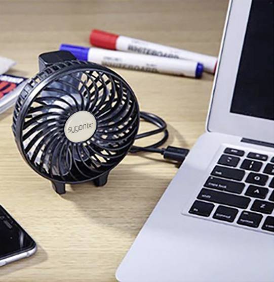 Mały czarny wentylator stołowy z złączem USB stoi obok laptopa. W tle na stole leżą dwa długopisy.