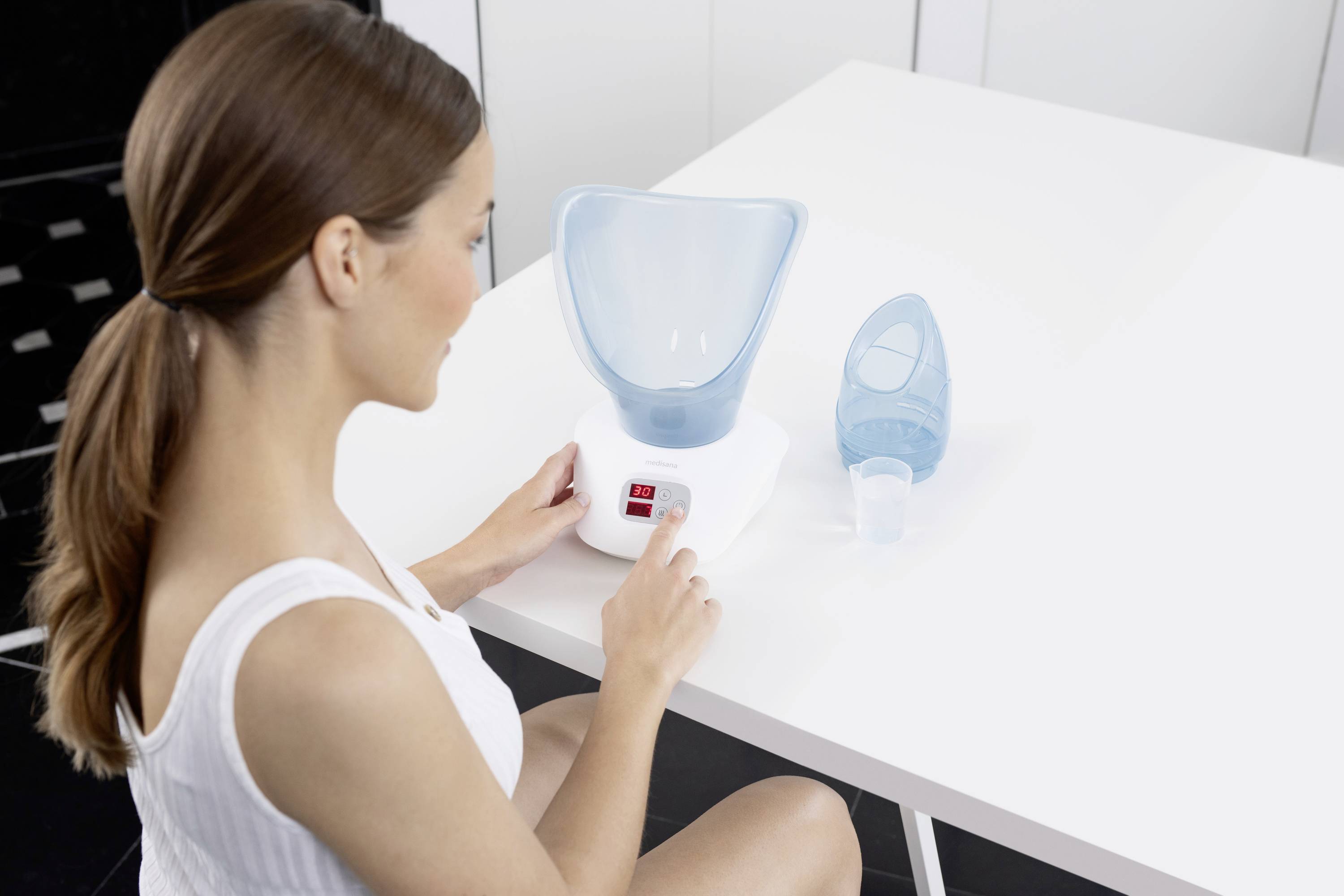 Kobieta obsługuje inhalator na stole. Obok niej stoi mniejszy pojemnik z wodą. Pomieszczenie jest urządzone w minimalistycznym stylu.