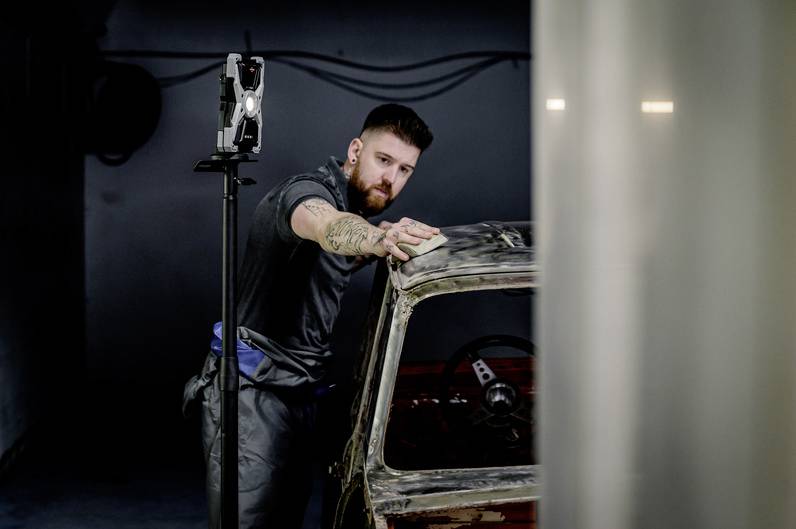 Mężczyzna poleruje stare auto szmatką w warsztacie. Na ramieniu ma wytatuowany wzór. W tle stoi lampa robocza.