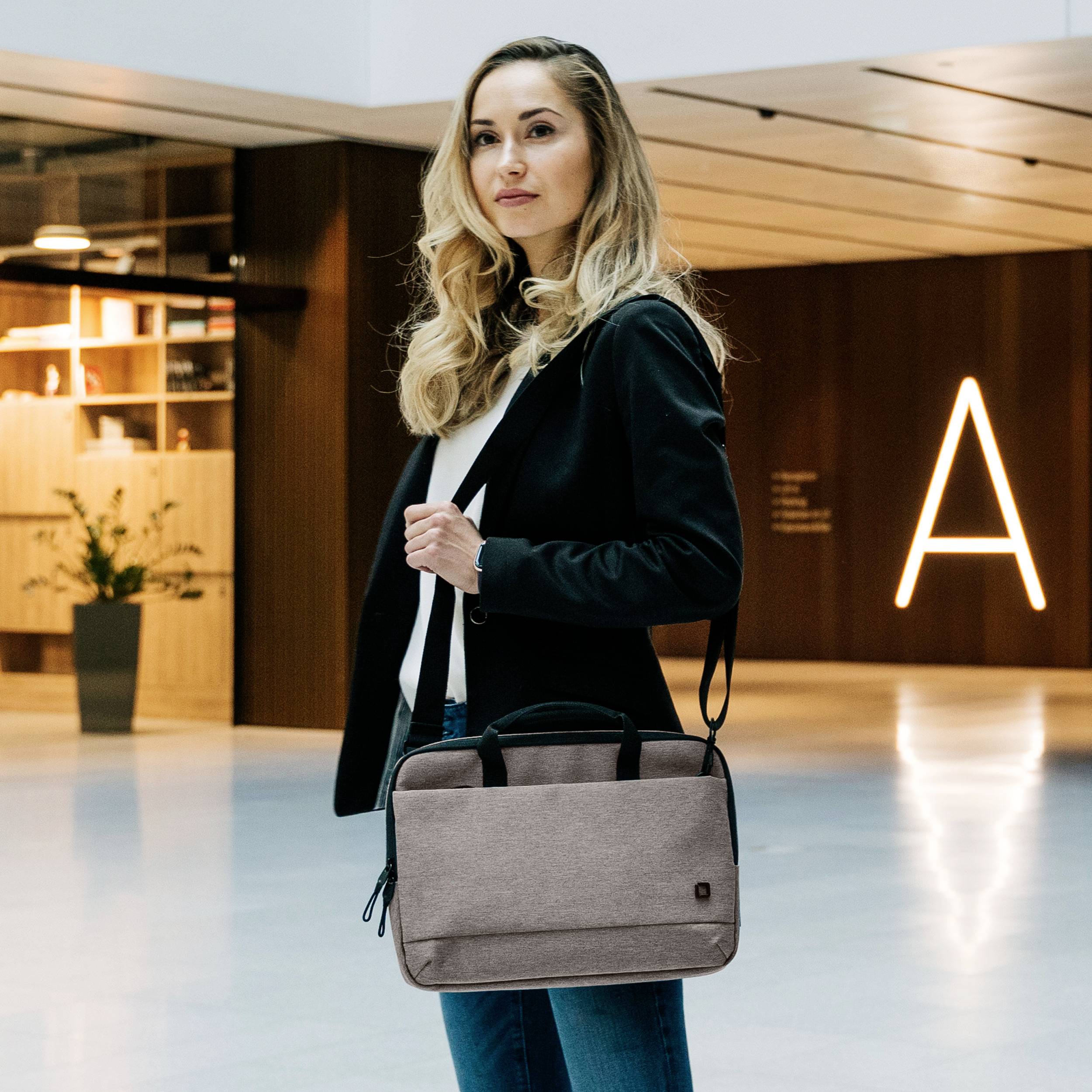 Osoba w nowoczesnym budynku, ubrana w elegancki strój, trzymająca torbę na laptopa. W tle stylowe drewniane ściany i nastrojowe oświetlenie.