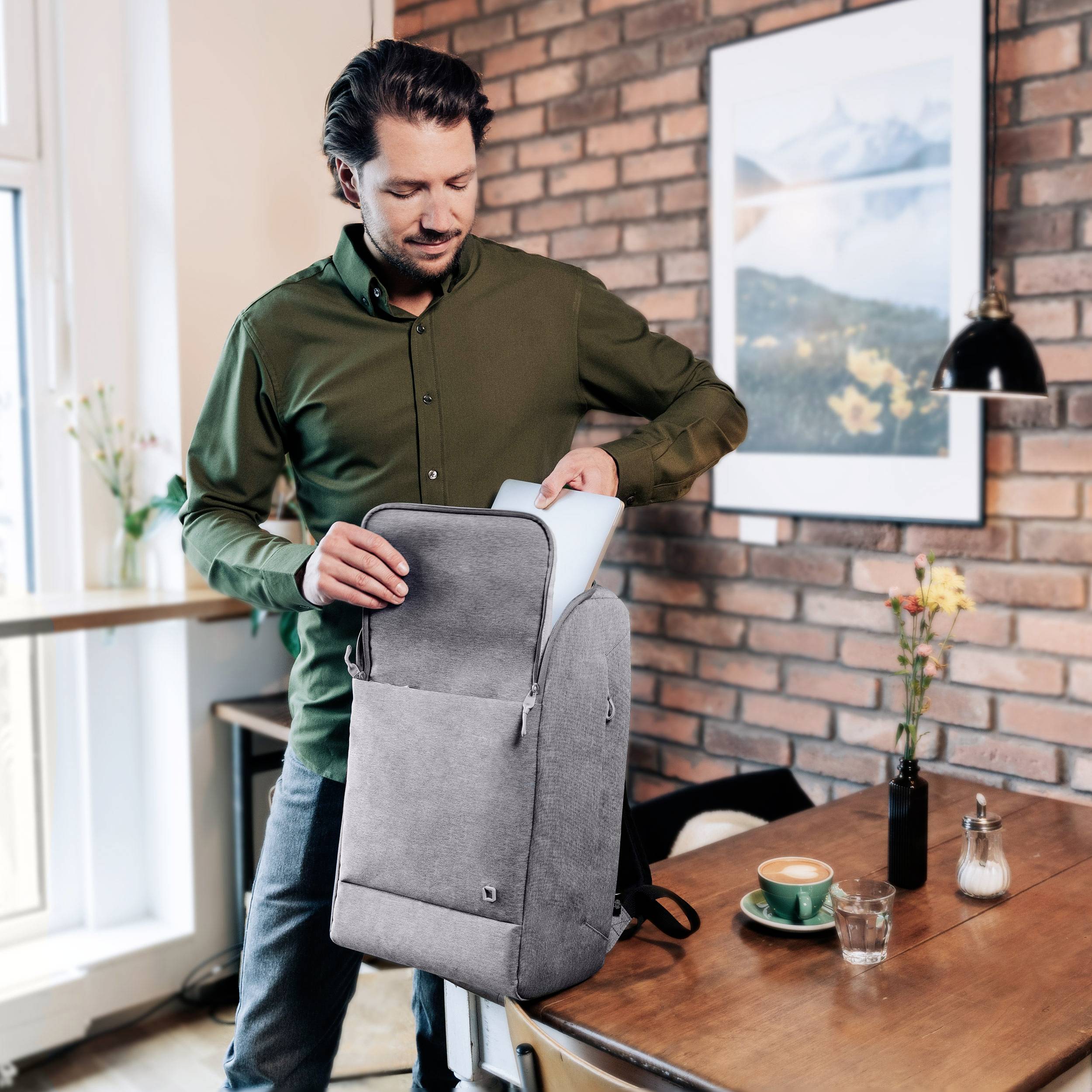 Mężczyzna wyjmuje laptopa z plecaka w kawiarni. Na stole stoją filiżanka kawy i talerz.