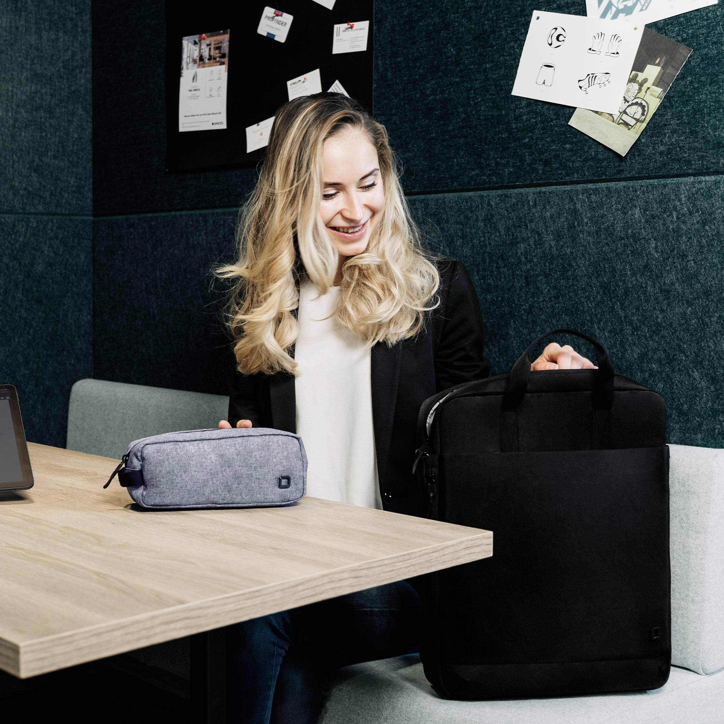 Uśmiechnięta kobieta siedzi przy stole i trzyma szarą torbę. Obok stoi czarna teczka. W tle widać tablicę informacyjną.