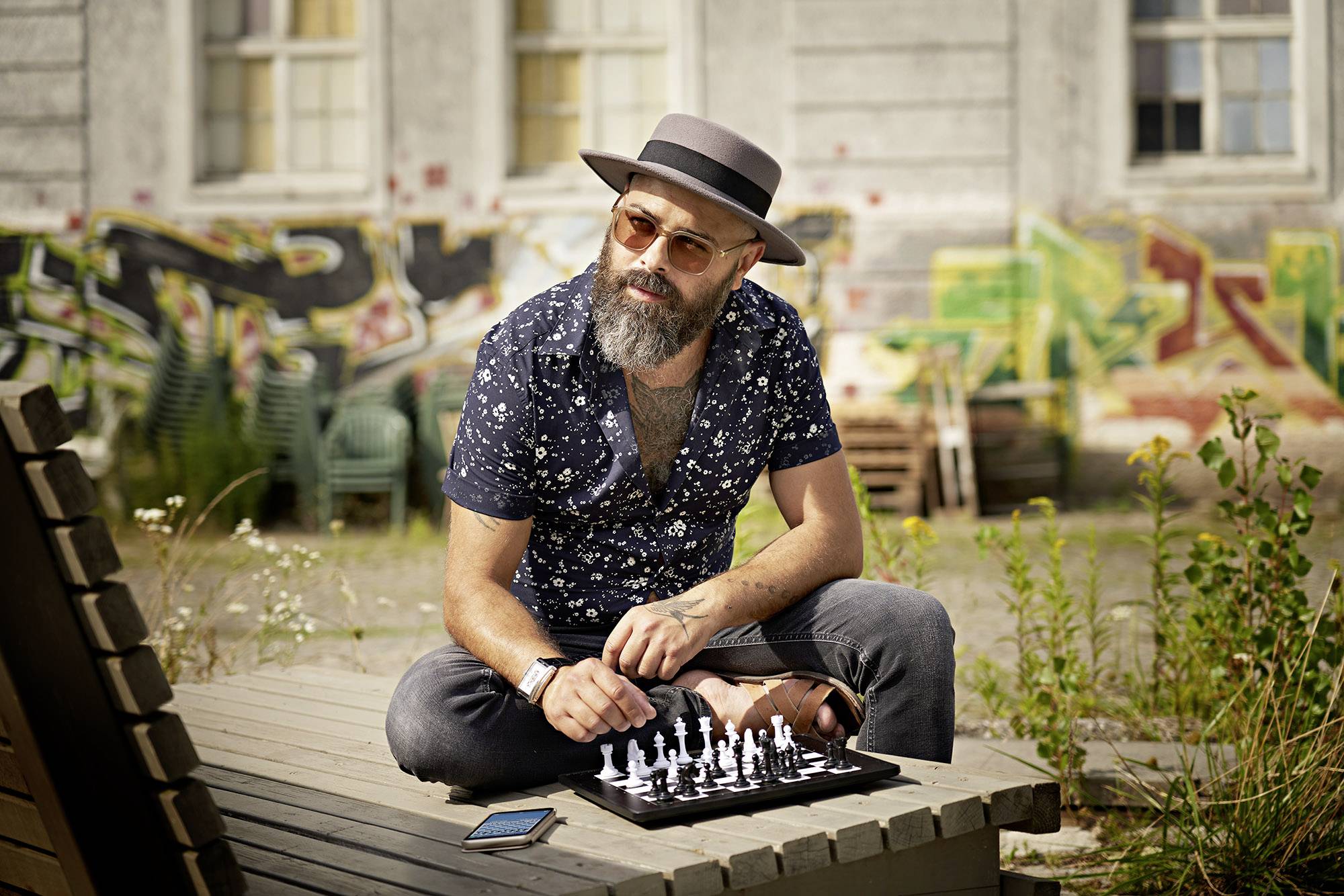 Mężczyzna w kapeluszu i okularach przeciwsłonecznych gra w szachy na wolnym powietrzu przed ścianą pokrytą graffiti. Obok niego, na stole, leży smartfon.