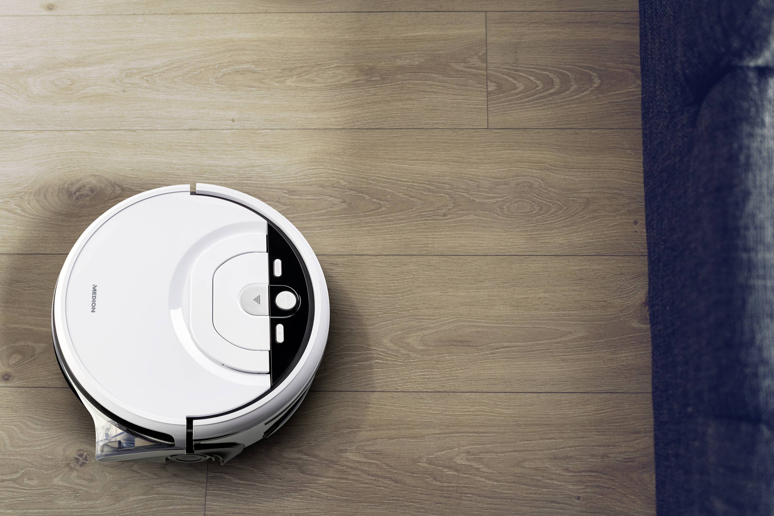 Biały robot sprzątający czyści drewnianą podłogę obok ciemnoszarej kanapy.