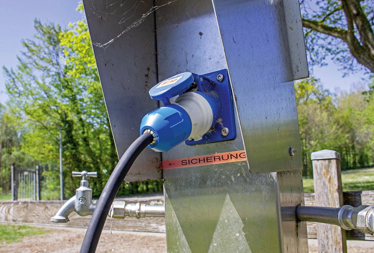 Zewnętrzne gniazdko elektryczne z podłączonym kablem zamontowane jest na metalowym słupie, obok kranu z wodą, w otoczeniu parku.