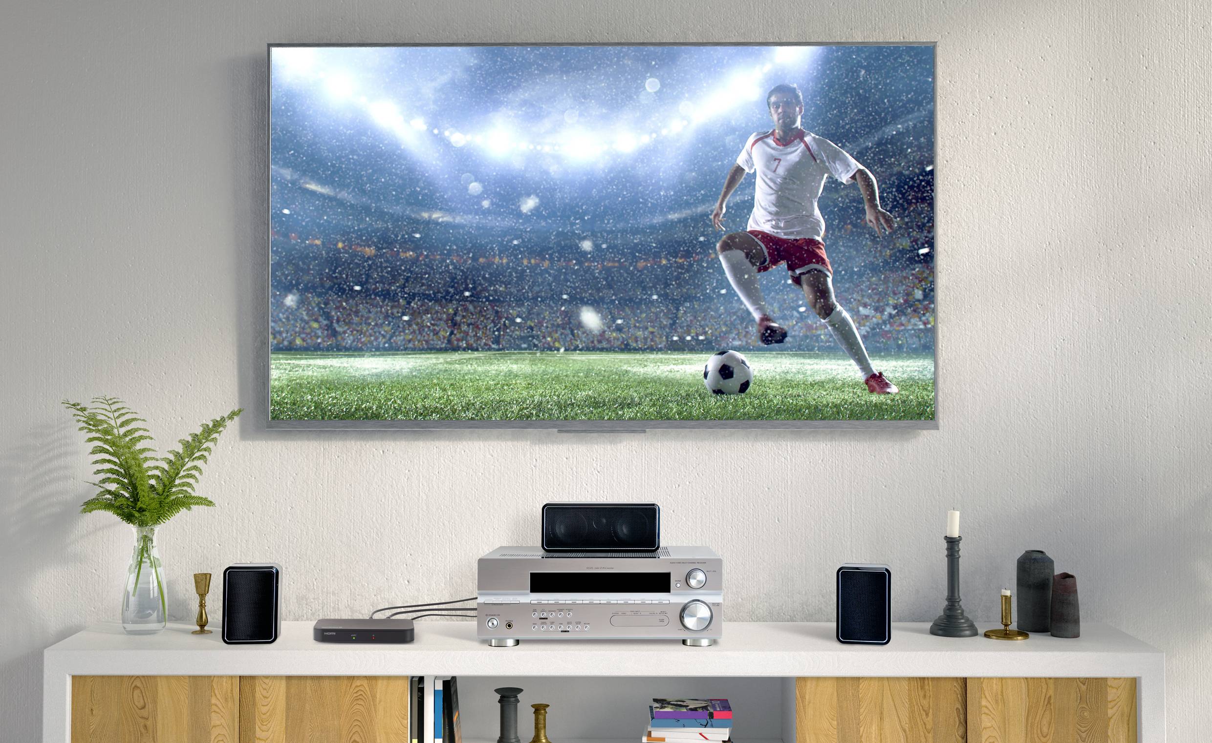 Salon z telewizorem, na którym wyświetlany jest mecz piłki nożnej. Zawodnik w czerwonych krótkich spodenkach prowadzi piłkę na oświetlonym stadionie.