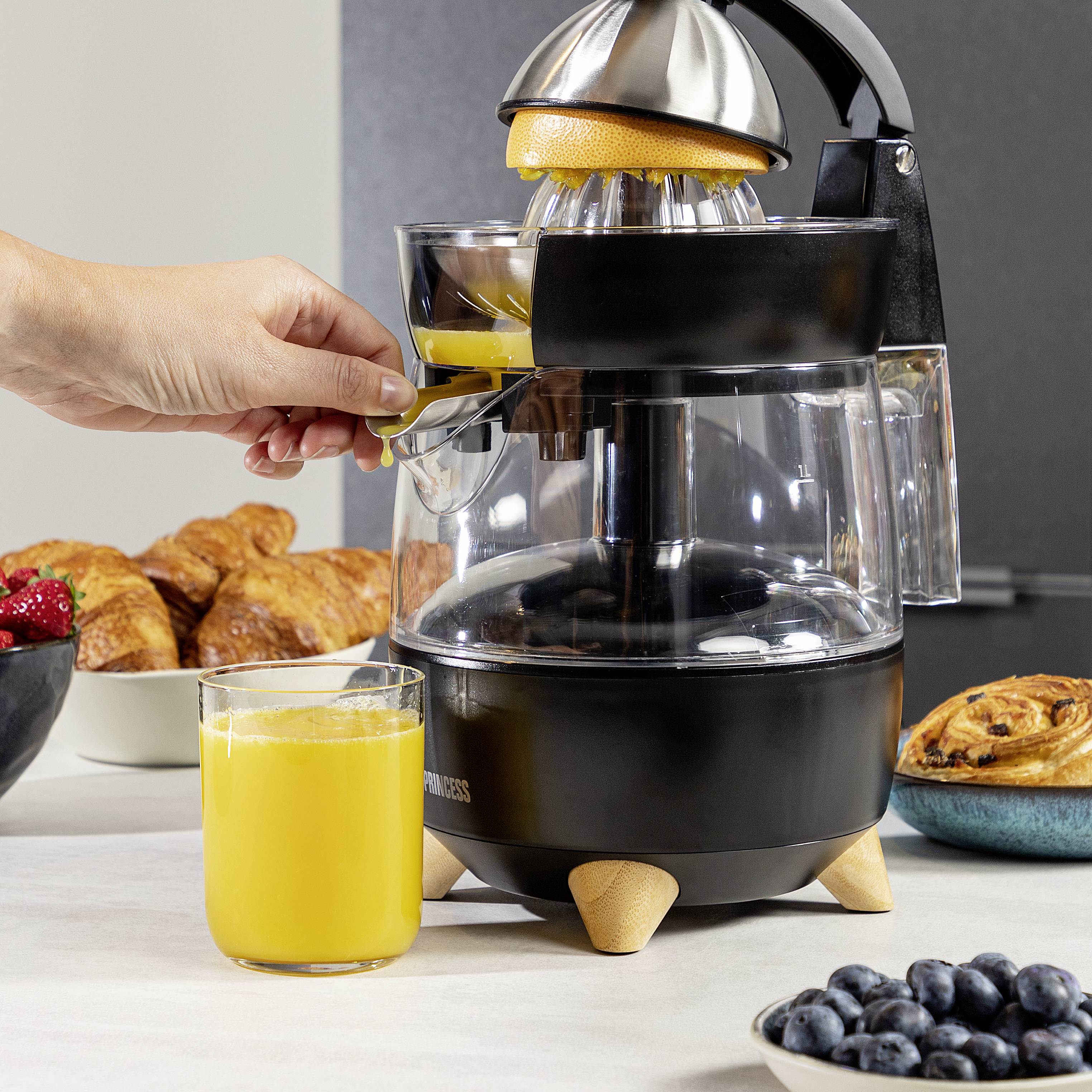Dłoń wyciska pomarańczę na wyciskarce do soków. Na pierwszym planie stoi szklanka świeżo wyciśniętego soku pomarańczowego. W tle widoczne są wypieki i owoce jagodowe.