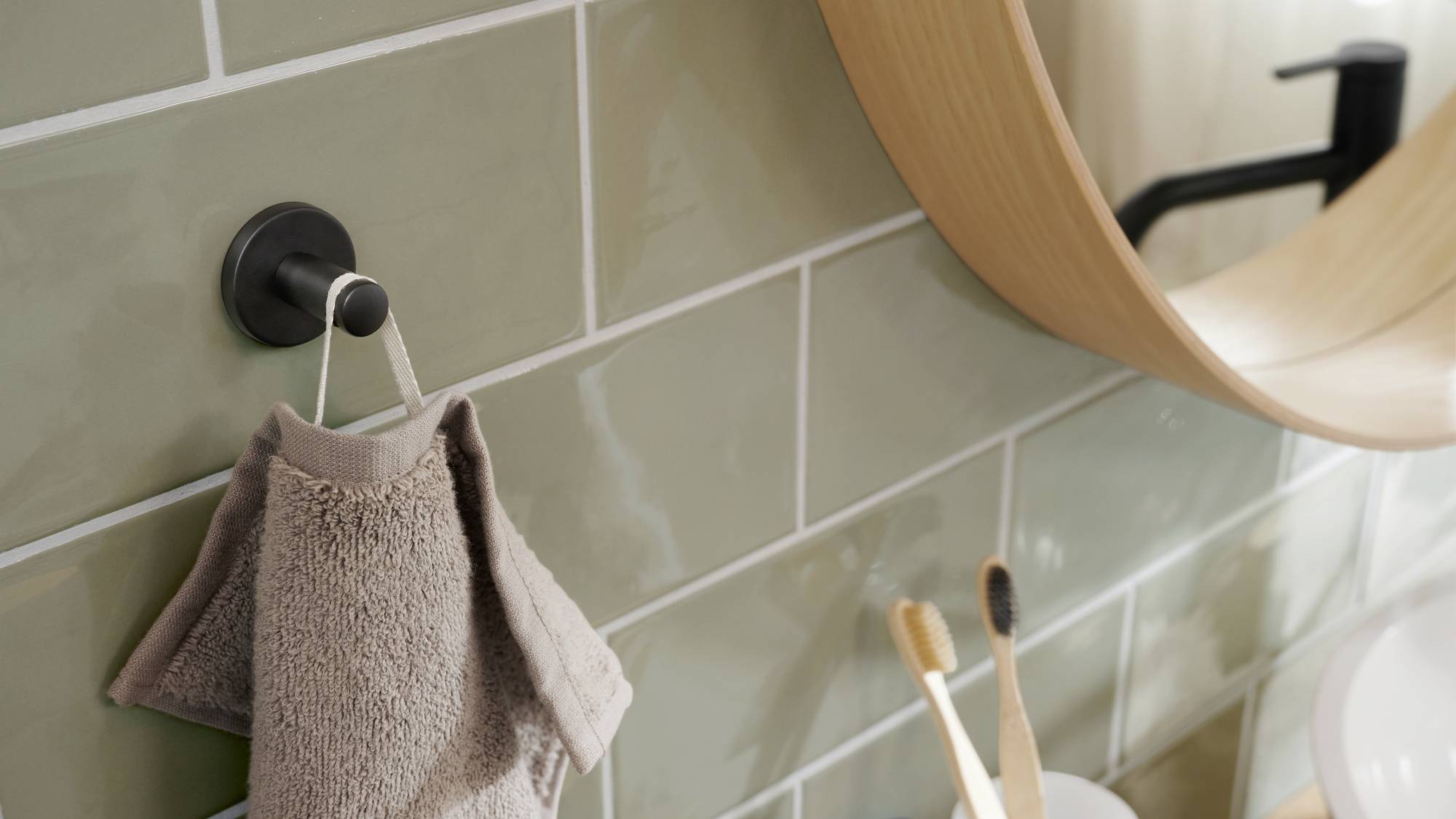 Ręcznik wisi na haczyku na kafelkowanej ścianie łazienki, obok okrągłe lustro, a na umywalce stoją dwie szczoteczki do zębów w uchwycie.