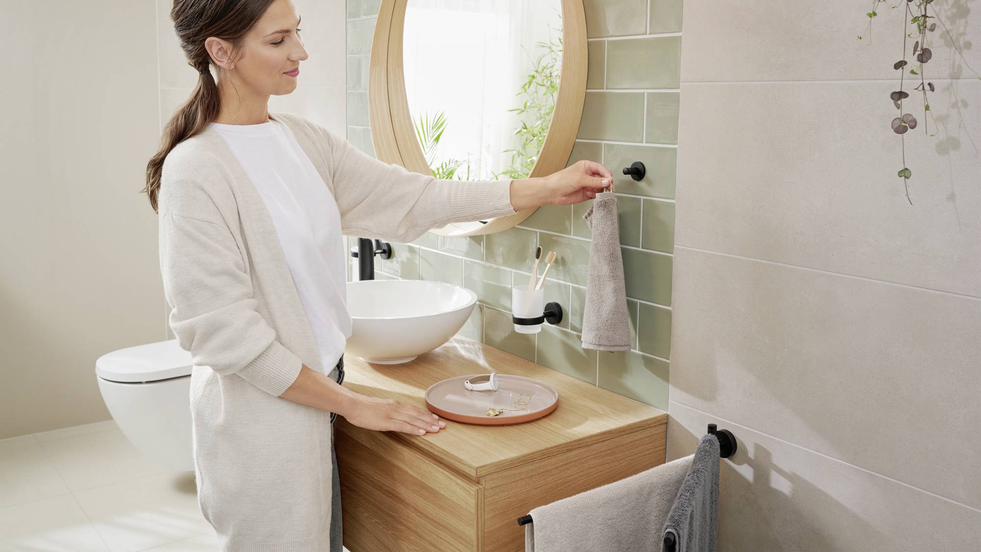 Kobieta w łazience zawiesza ręcznik na czarnym haku. Pomieszczenie jest nowocześnie urządzone, z okrągłym lustrem i zielonymi kafelkami.