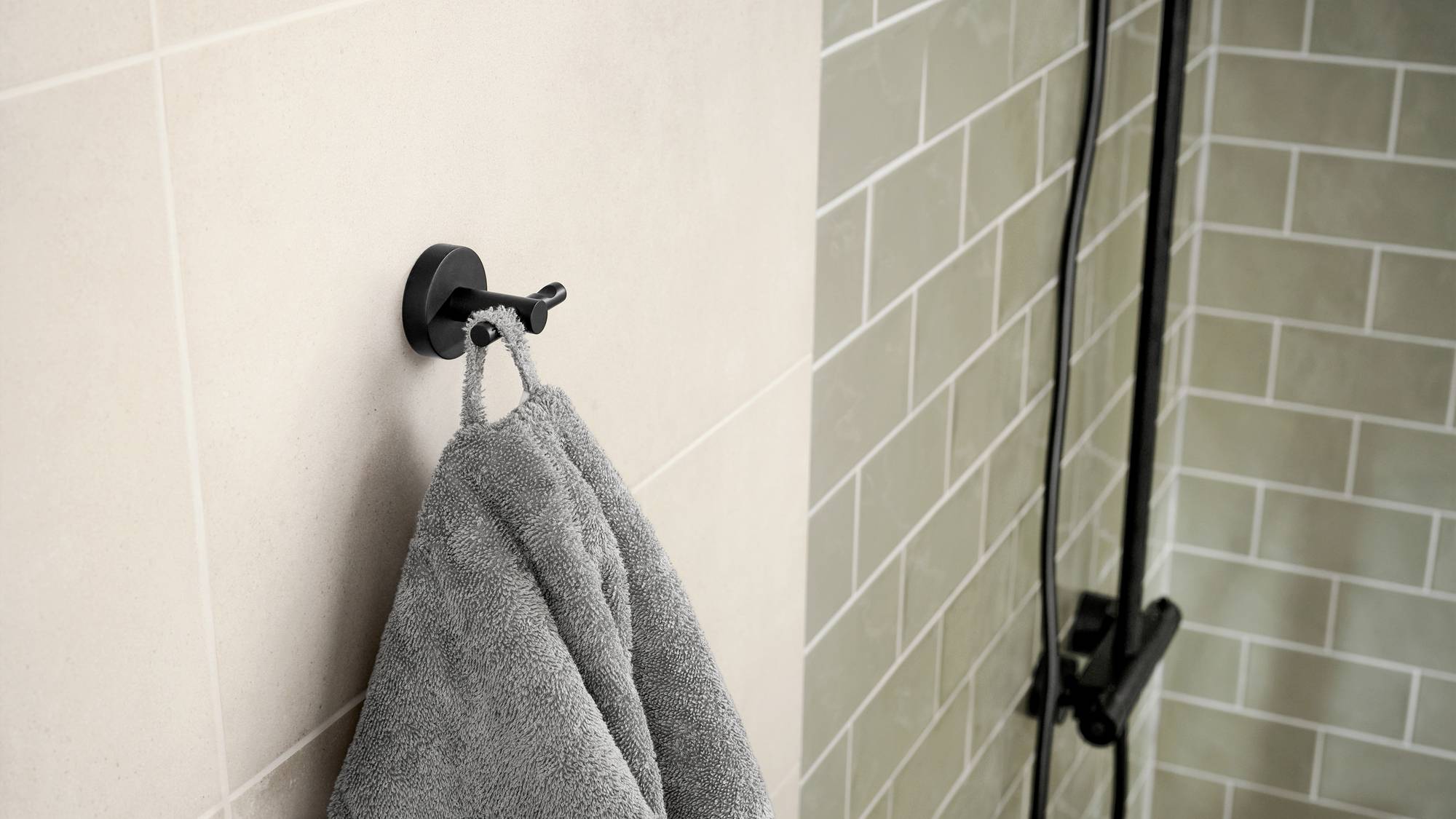 Szary ręcznik wisi na okrągłym, czarnym haku ściennym w łazience o ścianach wyłożonych kafelkami w odcieniach beżu i szarozieleni.