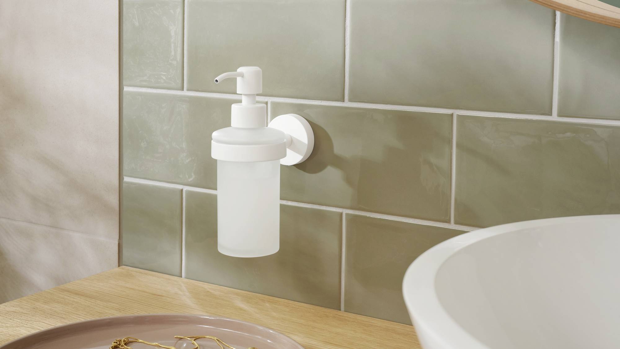 Prosty, biały dozownik do mydła jest zamontowany na ścianie wyłożonej zielonymi kafelkami, obok drewnianego blatu i umywalki.