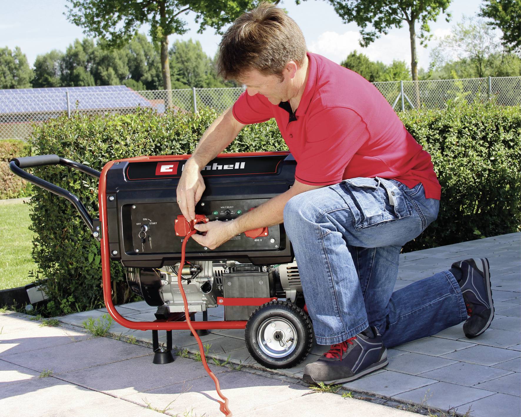 Mężczyzna w czerwonej koszuli klęczy na ziemi i obsługuje przenośny generator na świeżym powietrzu. W tle widoczne są drzewa i płot.