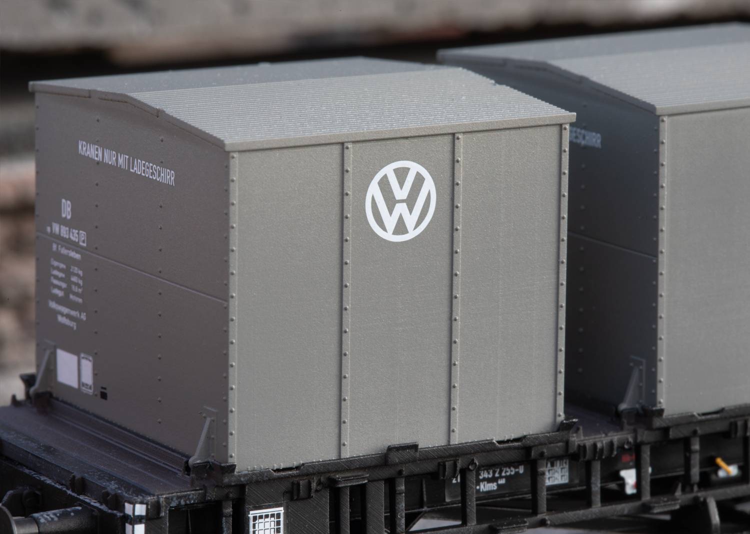 Szary wagon towarowy z logo VW na platformie kolejowej, prawdopodobnie przeznaczony do transportu części samochodowych lub komponentów.