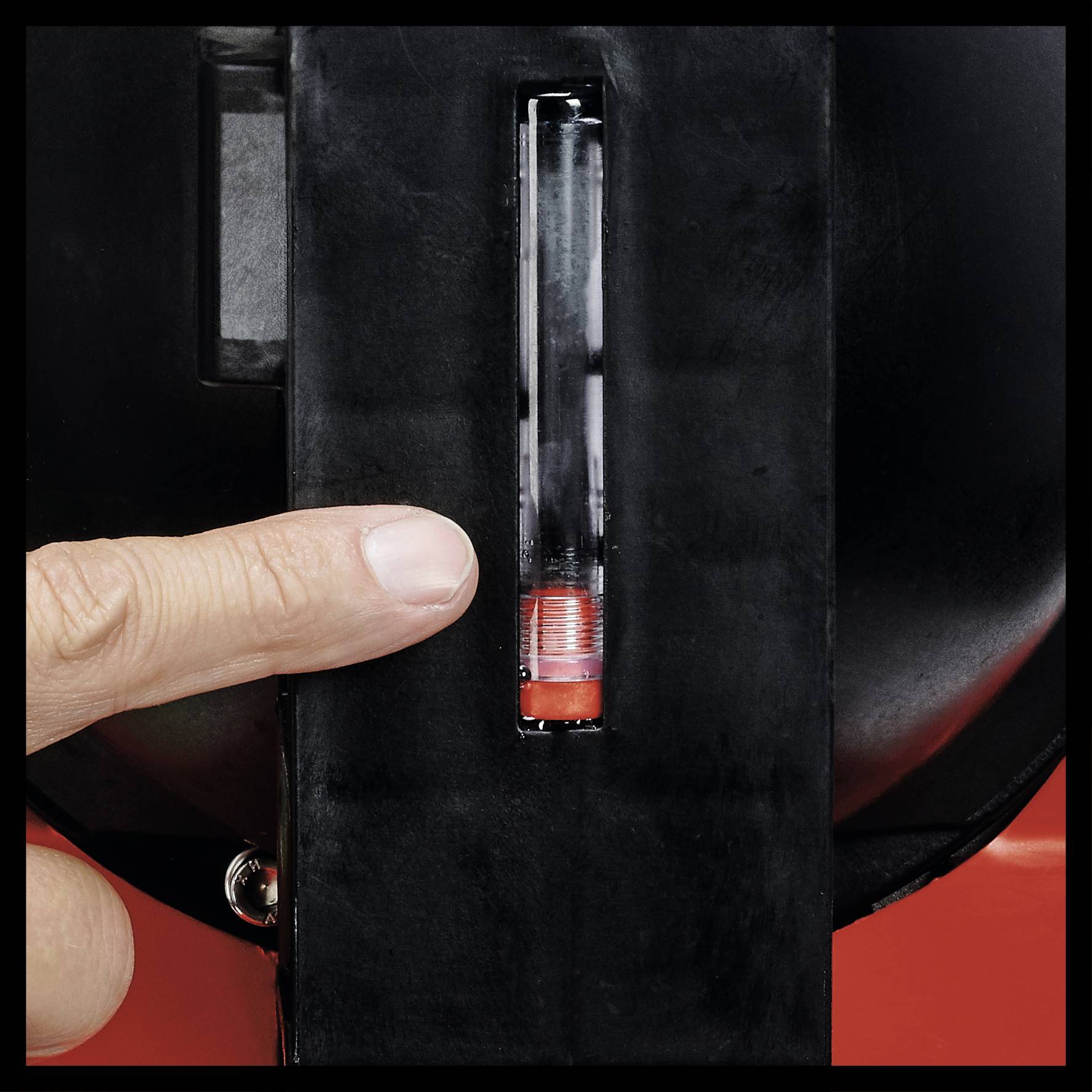 Palec wskazuje na przezroczysty cylinder z czerwonym tłokiem lub zaworem w czarnej obudowie.