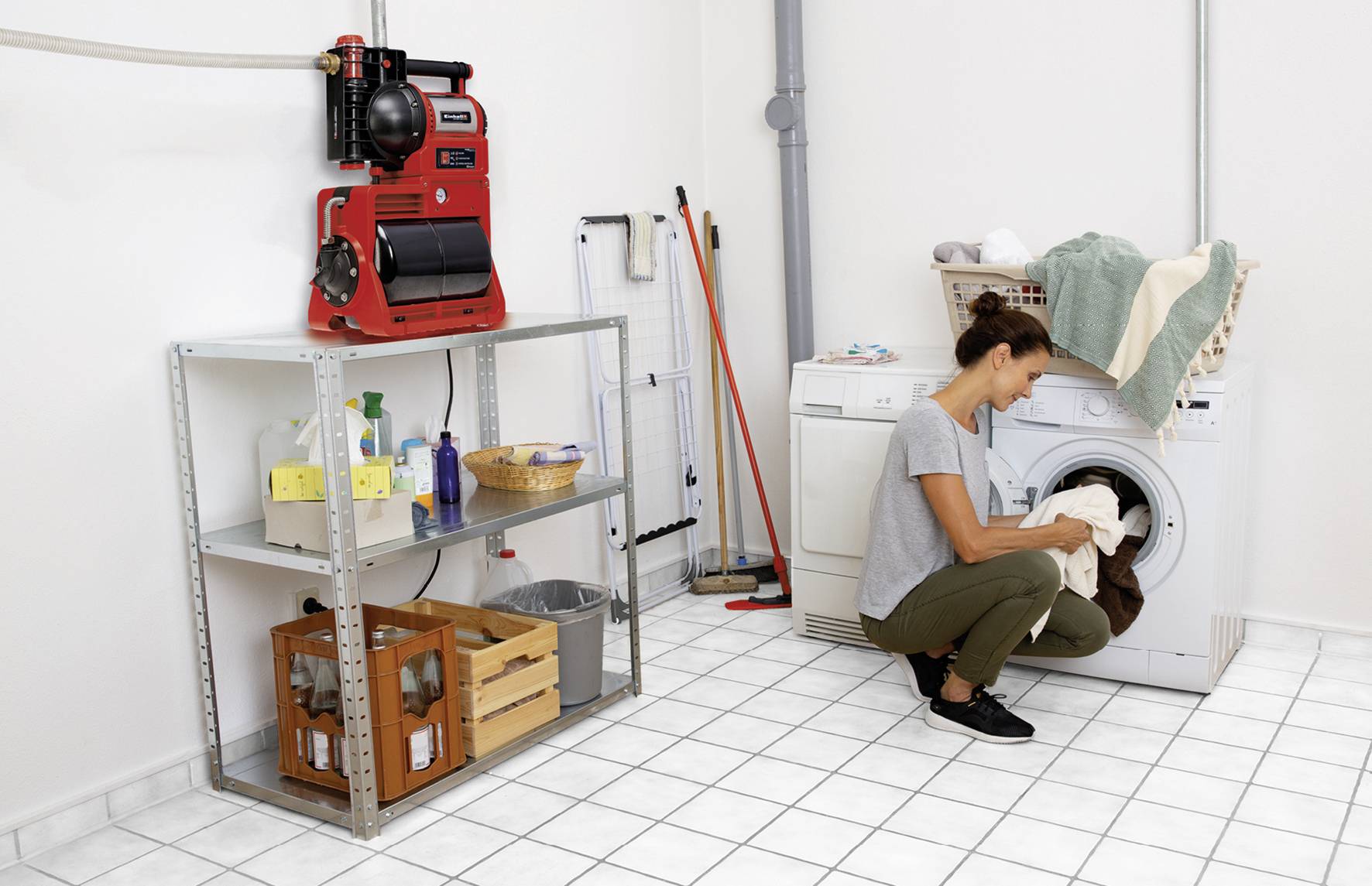 Kobieta ładuje pralkę w pomieszczeniu gospodarczym. Obok niej stoją środki czyszczące, a czerwony odkurzacz wisi na ścianie.
