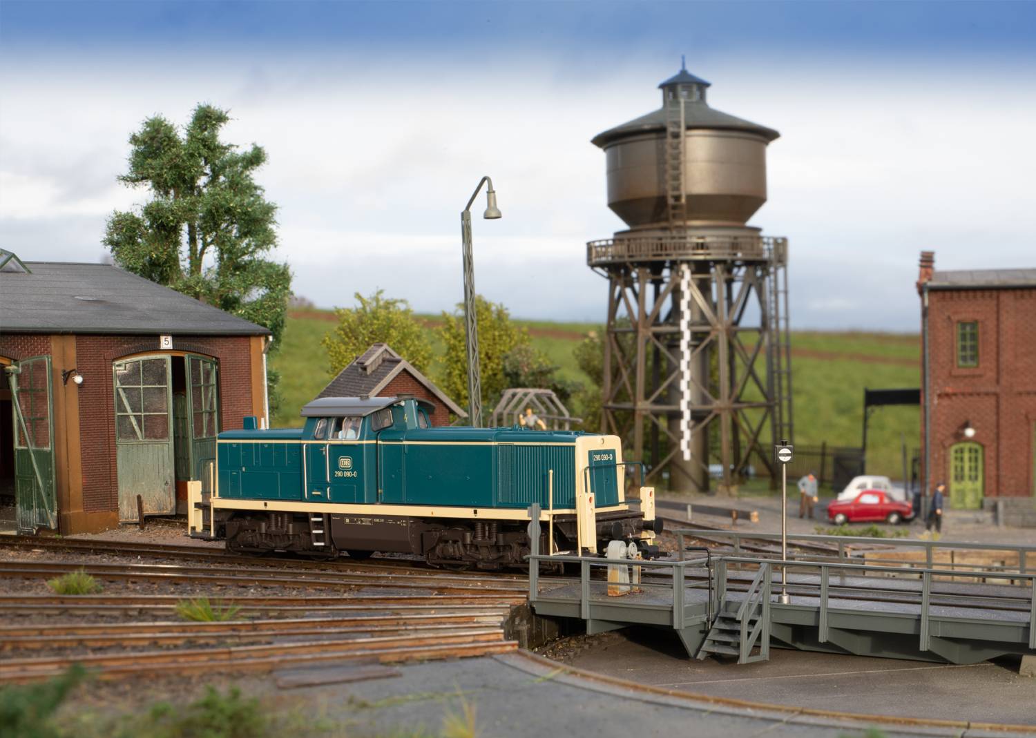 Sceneria makiety kolejowej z zieloną lokomotywą na torach, otoczoną budynkiem lokomotywowni, wieżą ciśnień i krajobrazem.