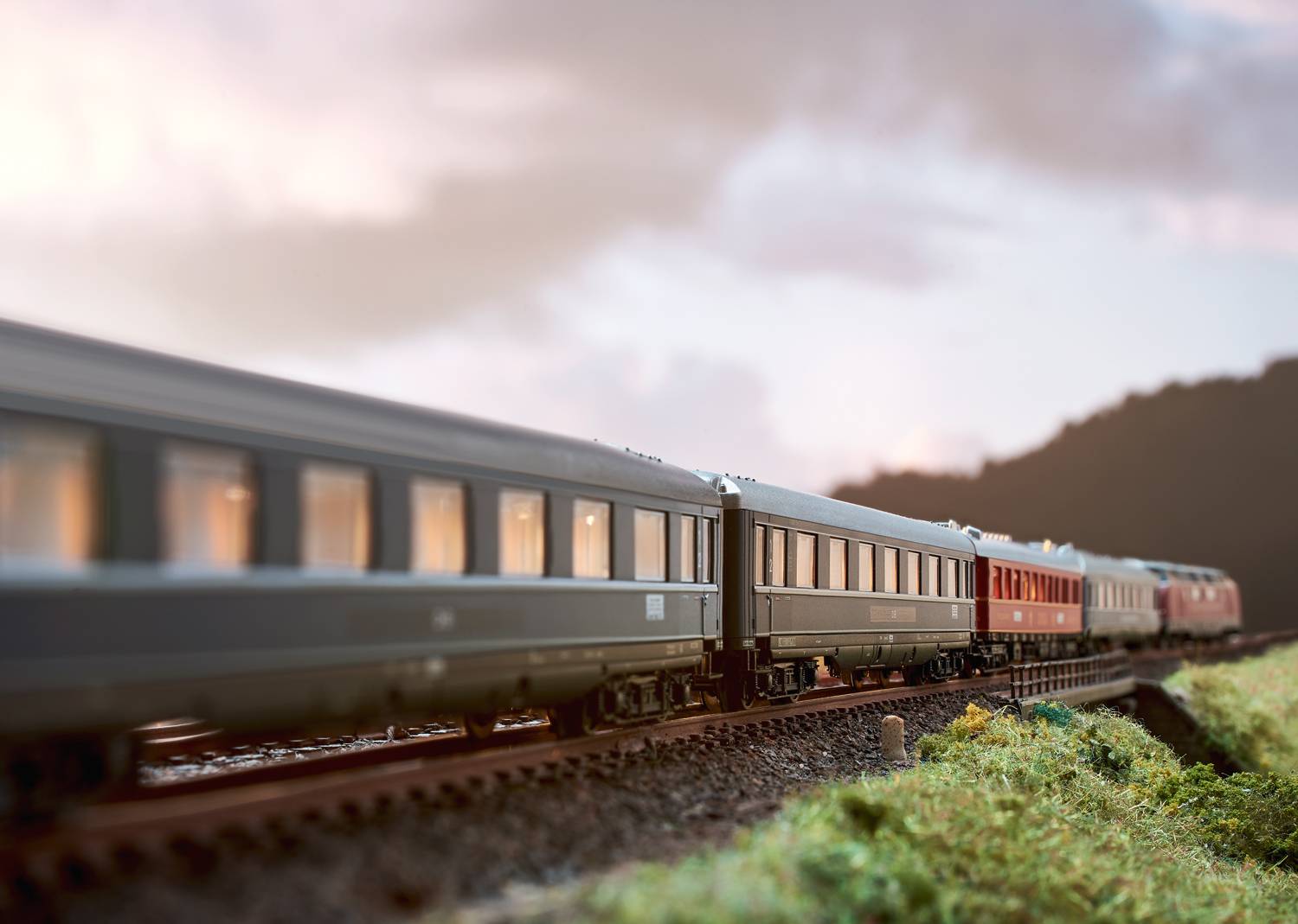 Pociąg na torach w wiejskim krajobrazie o zachodzie słońca. Niebo jest zachmurzone, pociąg jedzie od lewej do prawej strony.