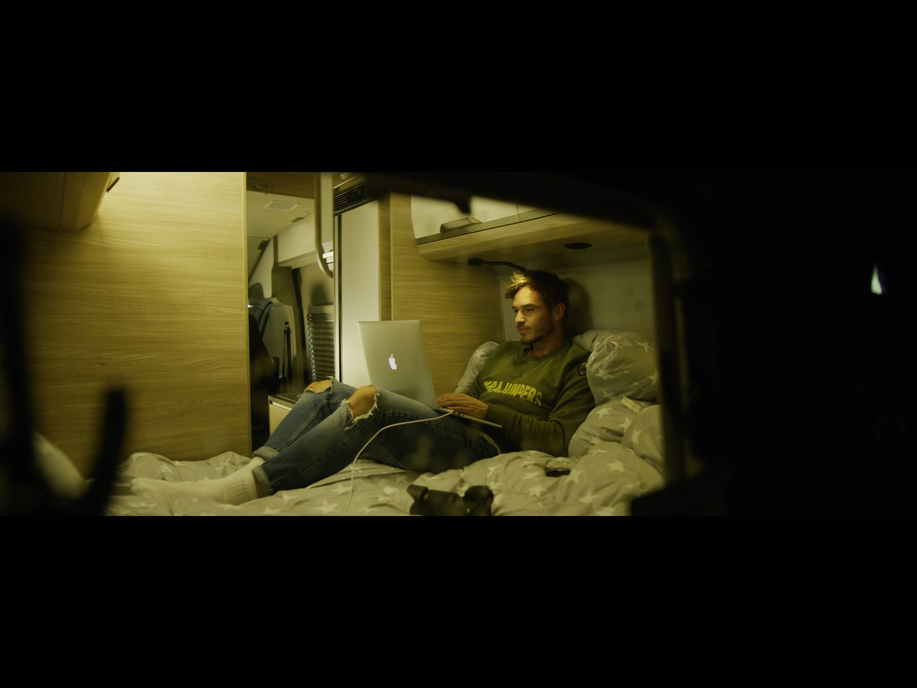 Osoba siedzi swobodnie na łóżku w kamperze i pracuje na laptopie.